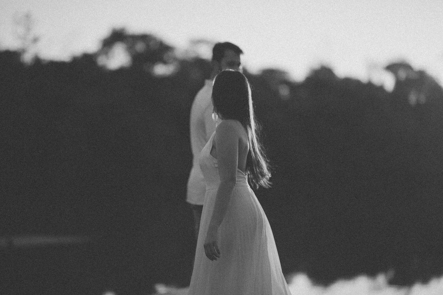 Black and white photo of couple in white attire standing by a calm lake.
