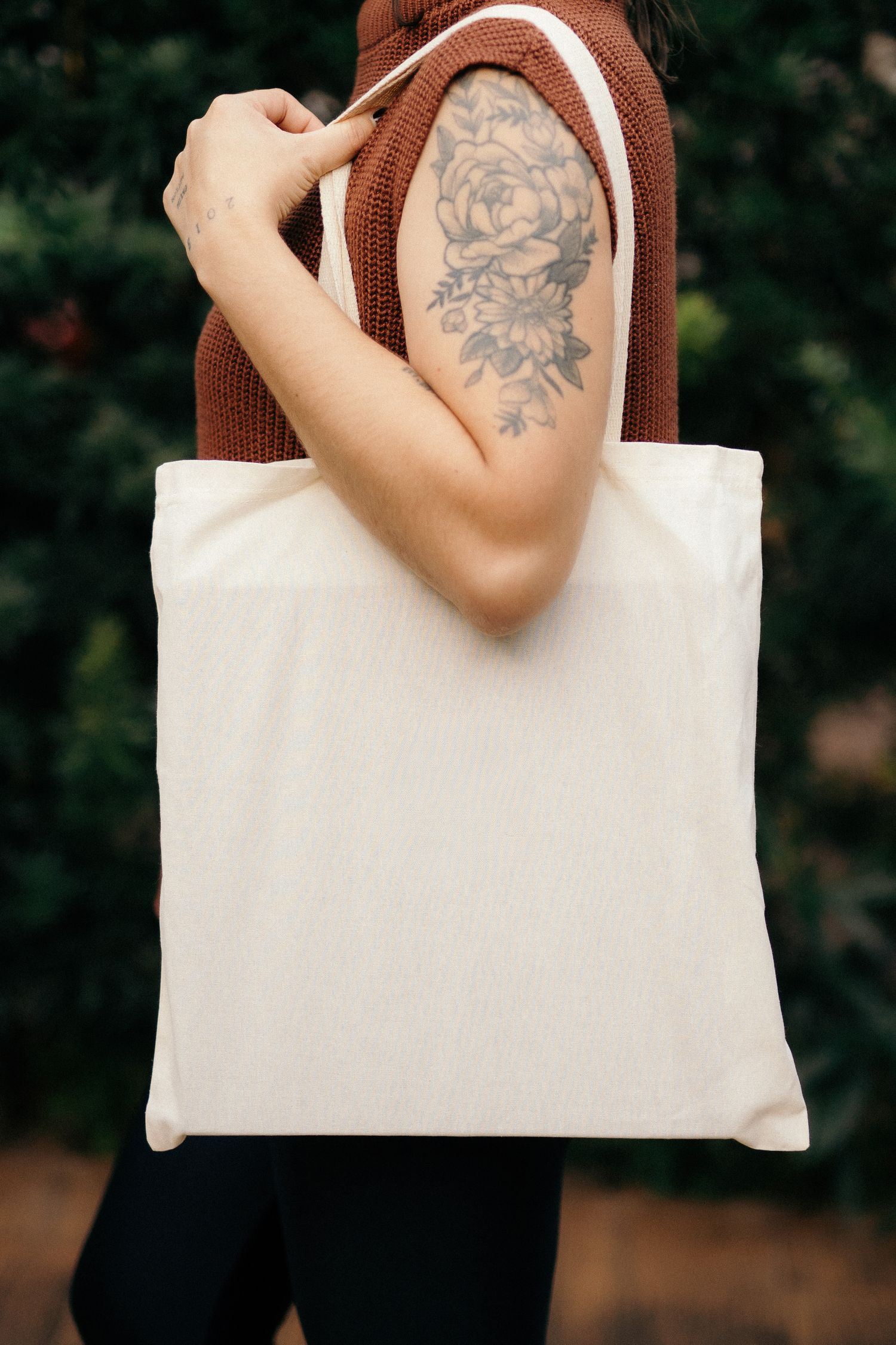 Woman with floral arm tattoo holding a blank white canvas tote bag outdoors.