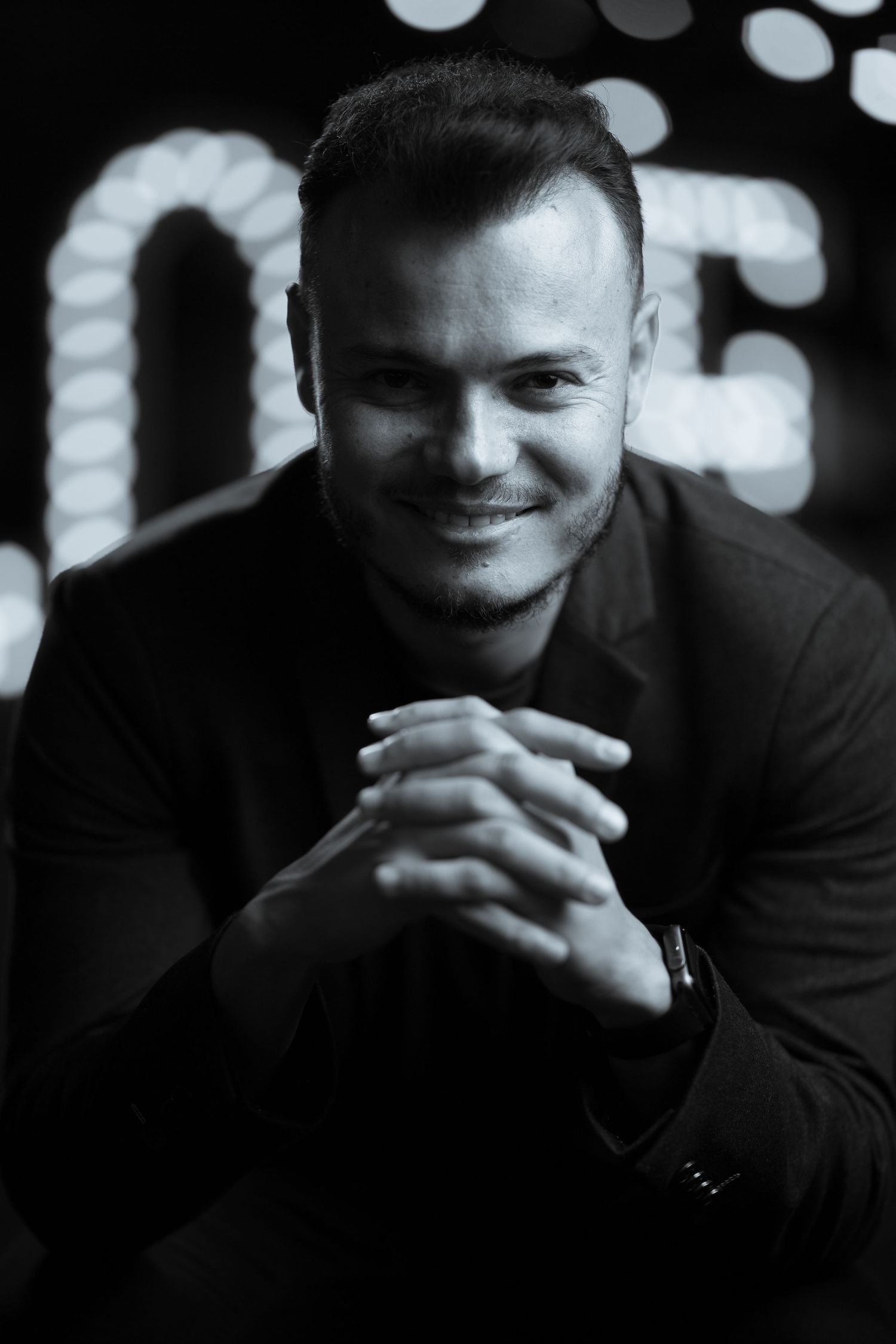 Black and white portrait of a smiling man in a dark suit with clasped hands against bokeh lights.