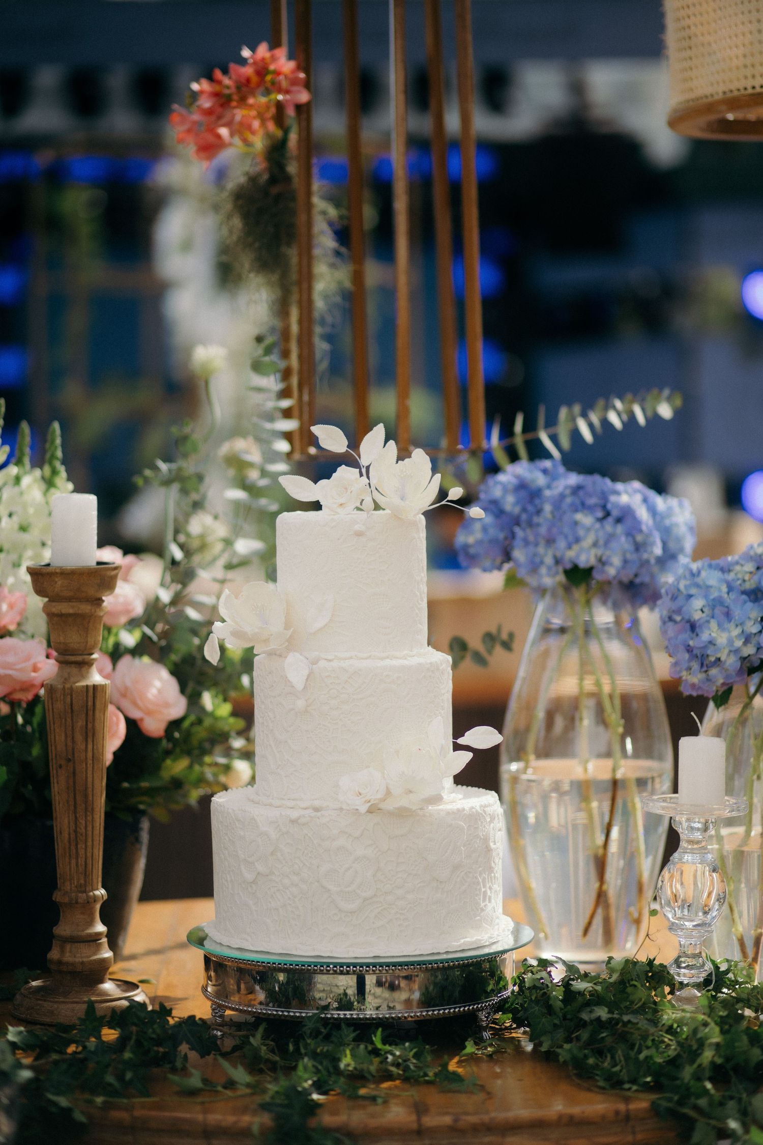 Elegant three-tier white wedding cake with floral decorations, surrounded by candles and blue hydrangeas on a decorated table.
