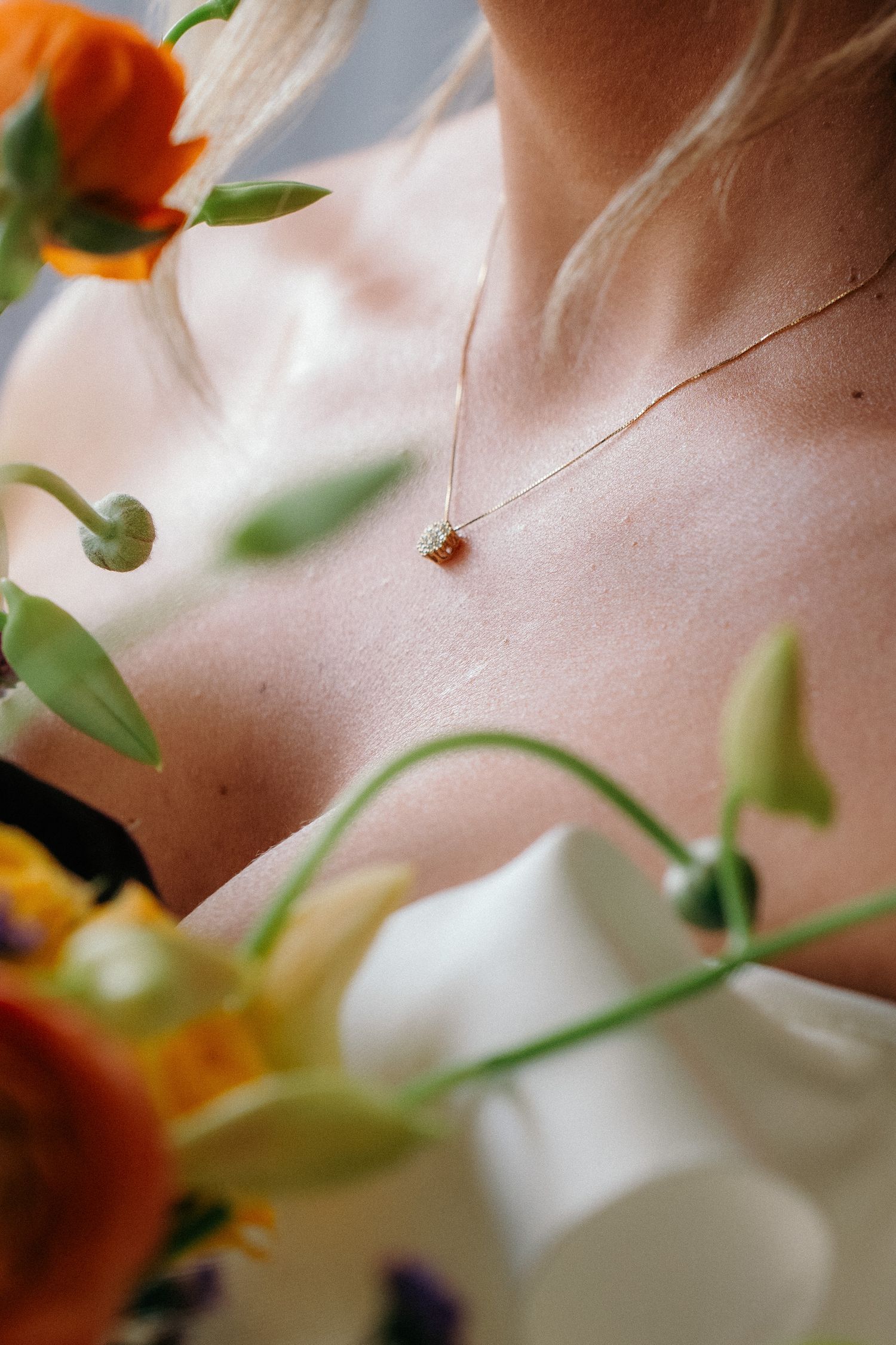 Close-up of bride wearing delicate gold pendant necklace surrounded by vibrant orange and white flowers.