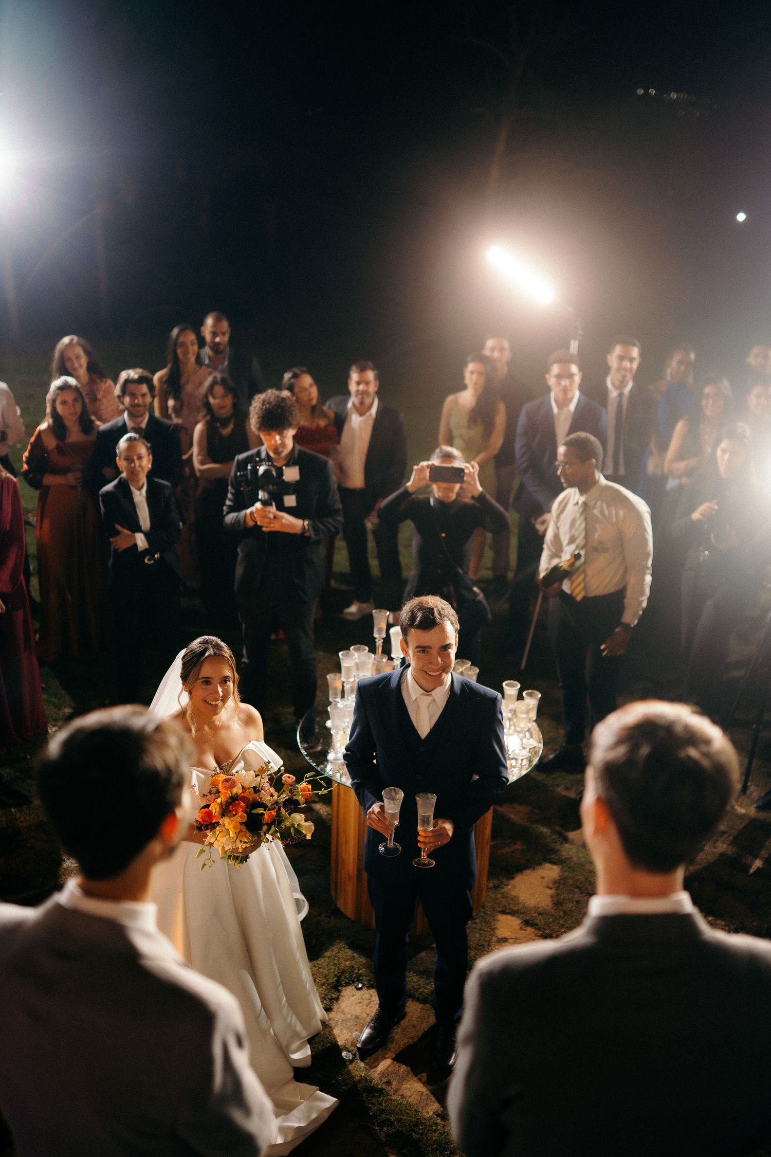 Aerial view of bride and groom seated at wedding reception surrounded by guests under bright lights at night.