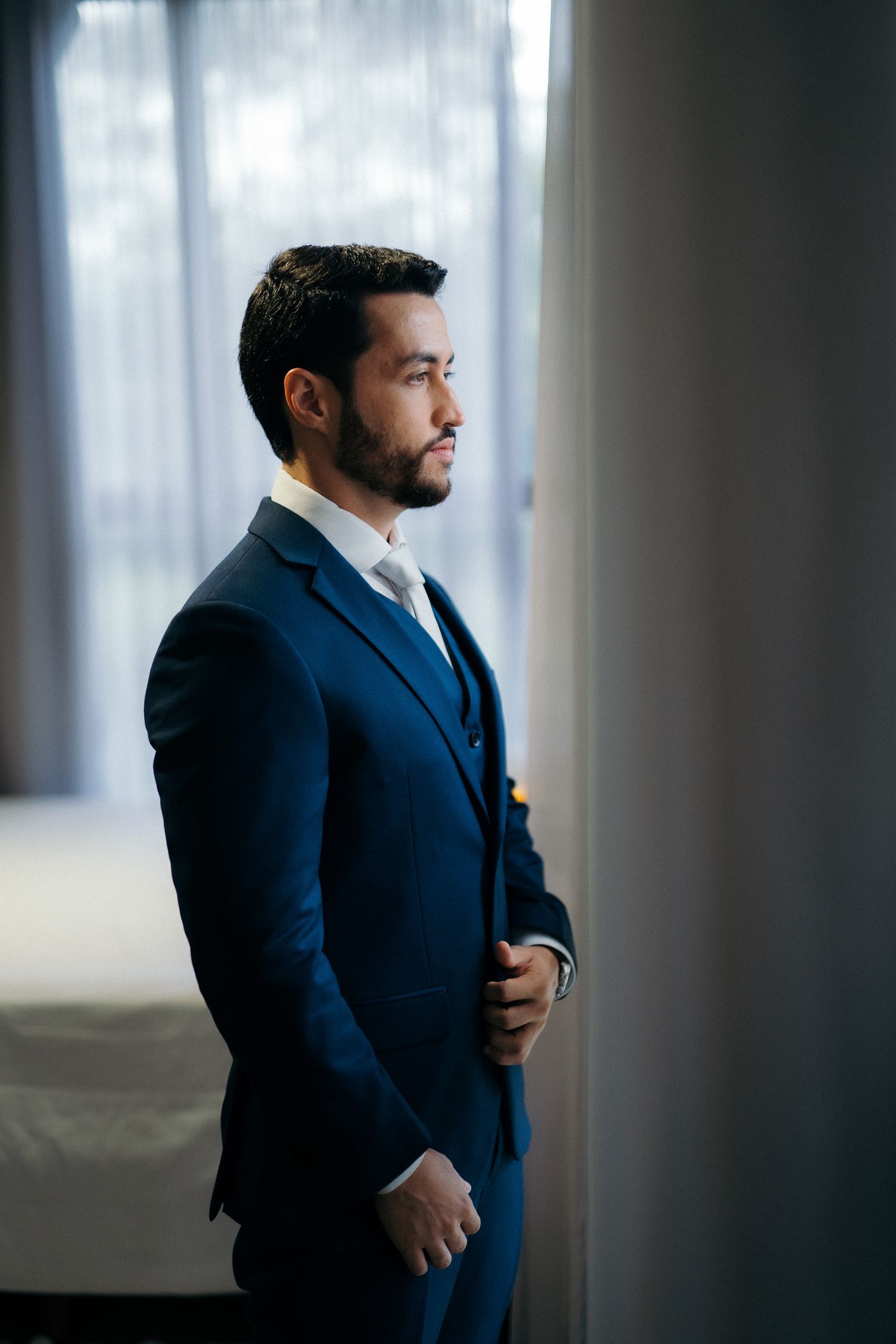 Groom in navy blue suit standing by window in hotel room before wedding ceremony.