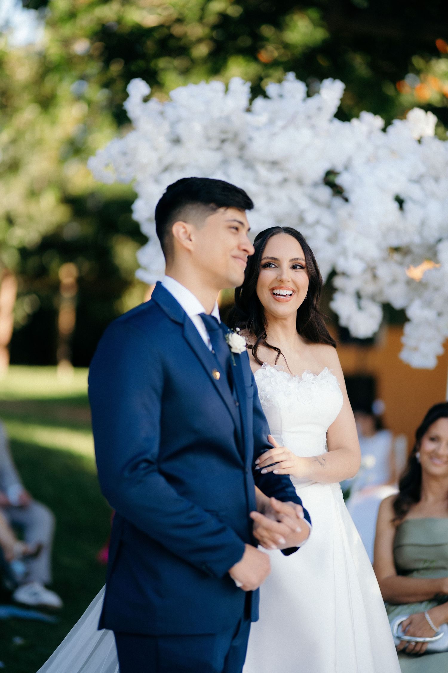 Bride and groom laughing joyfully at outdoor wedding ceremony with white floral arch backdrop.