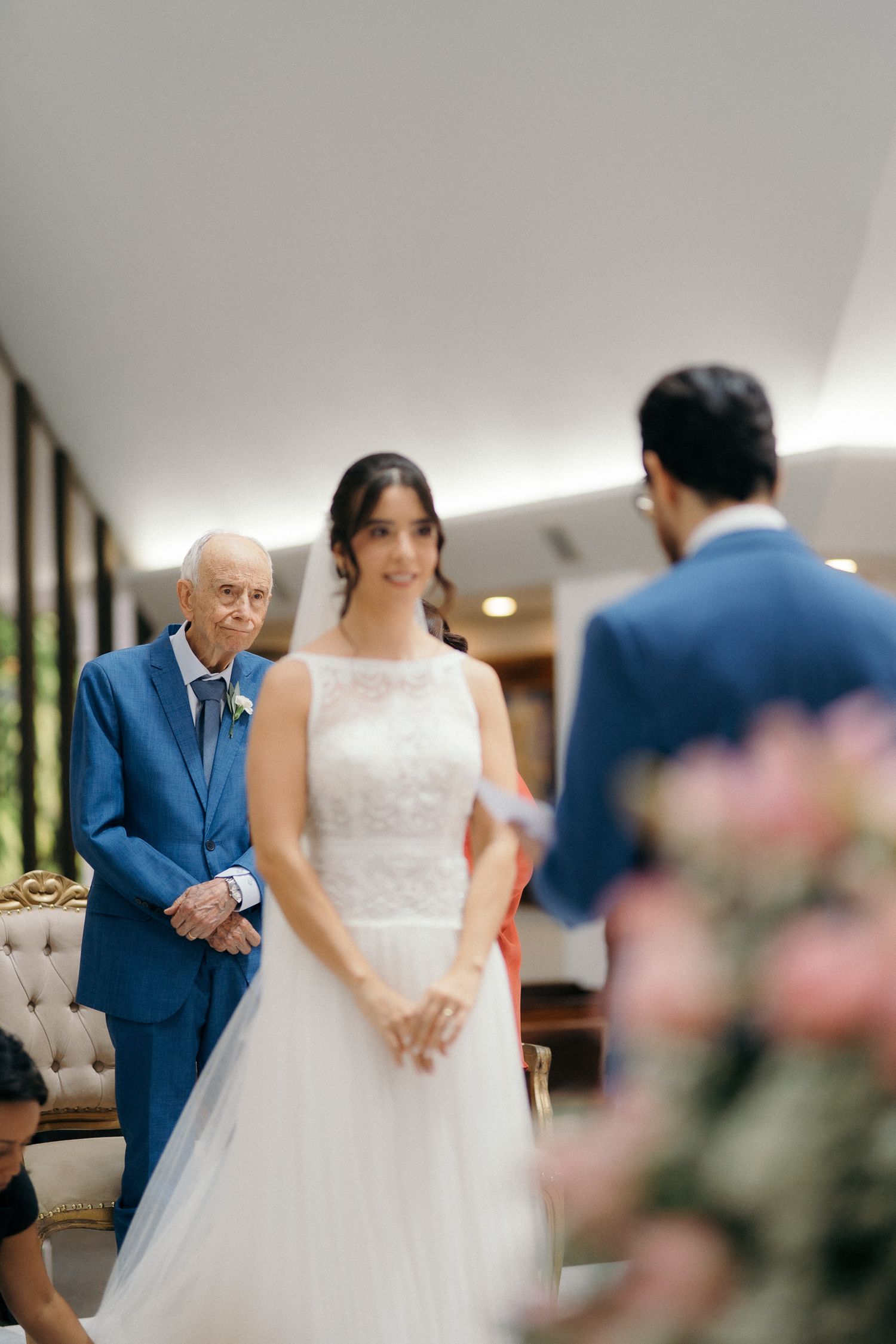 Bride in white lace gown stands facing groom during intimate indoor wedding ceremony with elderly guest seated behind.