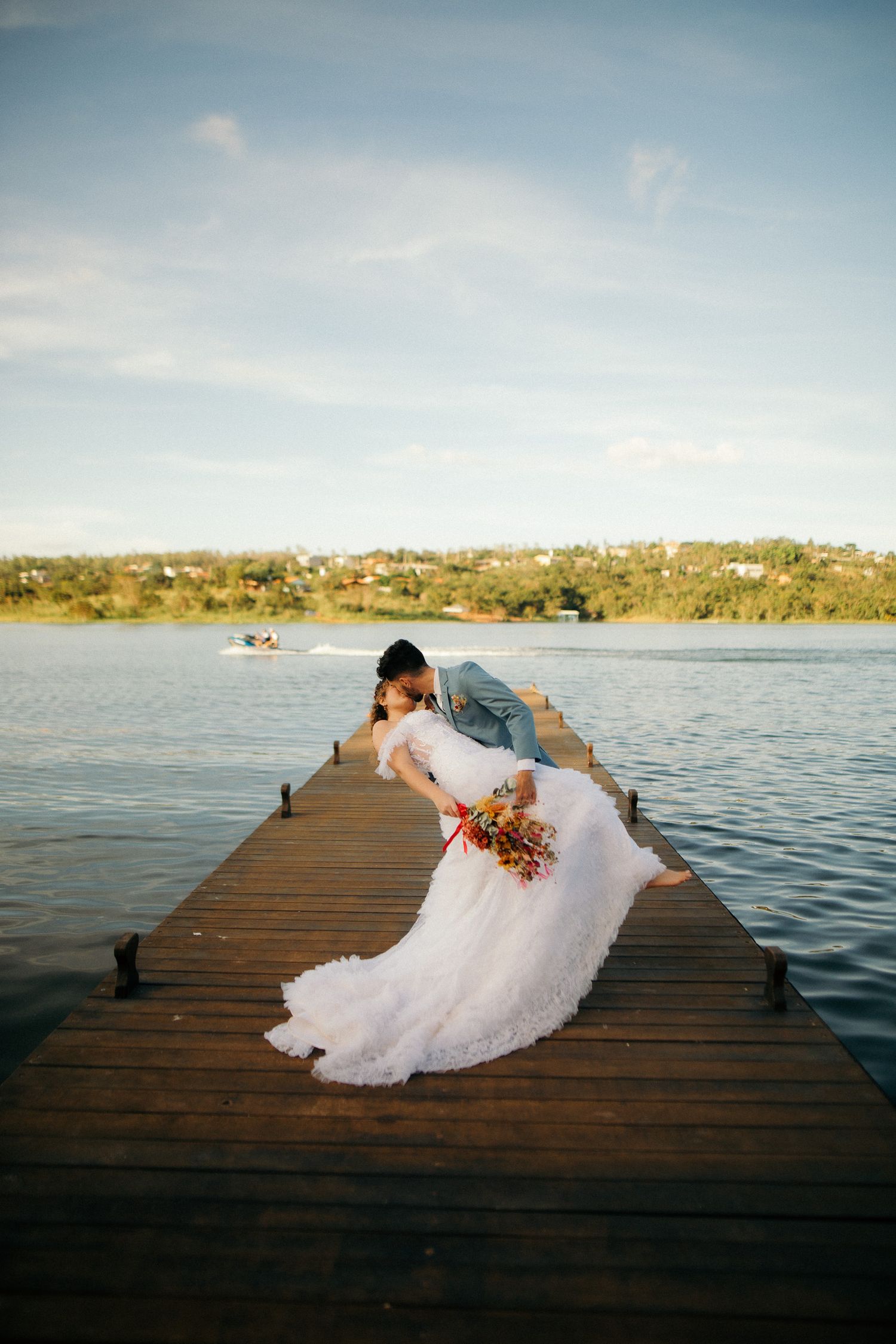 Bride and groom share a romantic dip kiss on a wooden pier overlooking a serene lake.