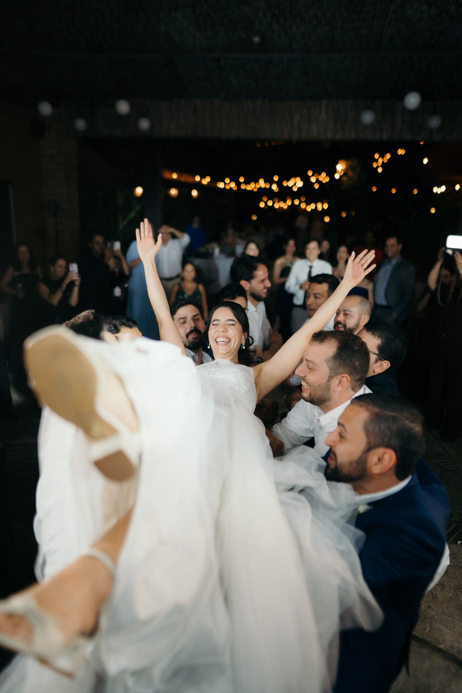 Bride laughing joyfully while being lifted by wedding guests during lively reception dance.