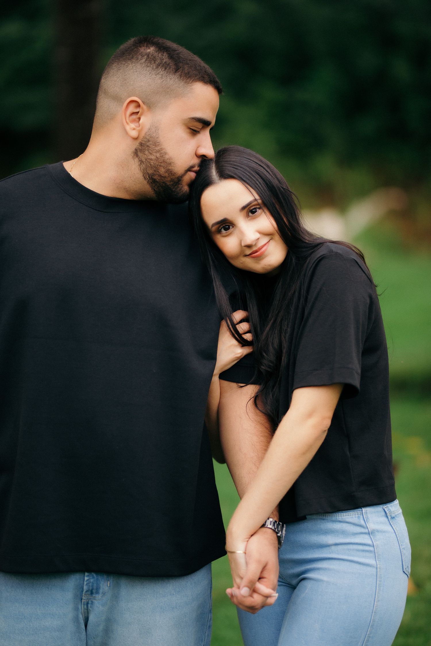 Couple in matching black shirts and jeans holding hands tenderly in a lush green outdoor setting.
