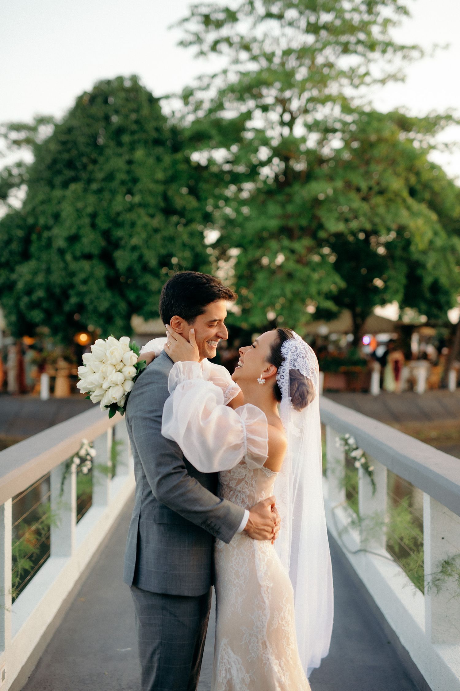 Smiling bride and groom embrace on white bridge surrounded by lush green trees.