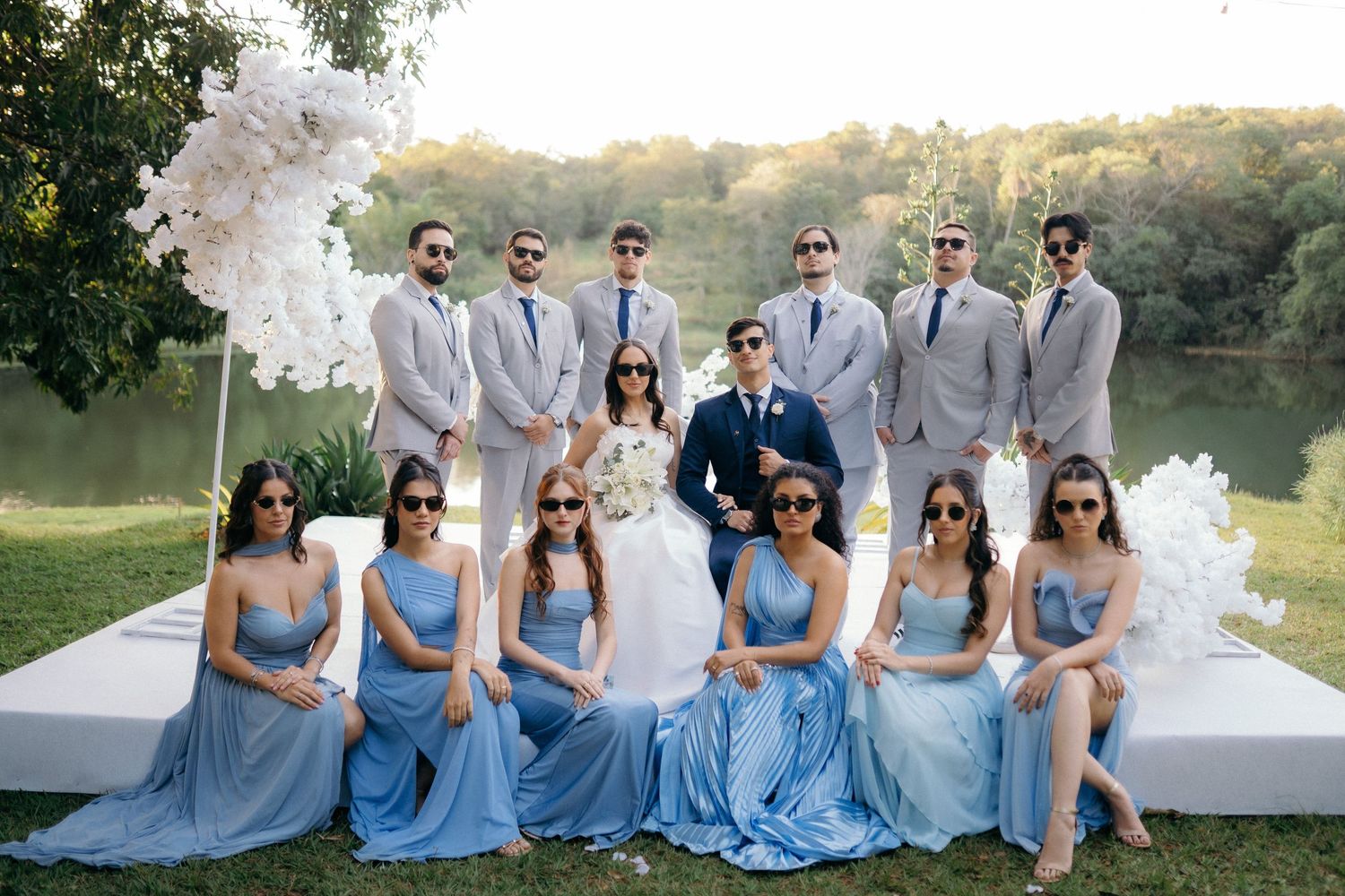 Wedding party poses with bride and groom, groomsmen in gray suits and bridesmaids in blue dresses outdoors.