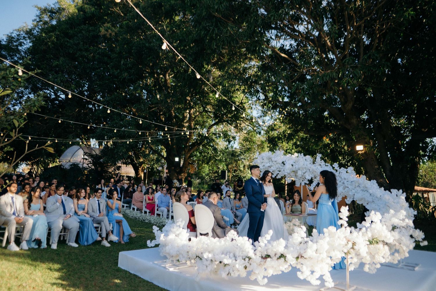 Outdoor wedding ceremony with white floral arch, guests seated on lawn, string lights, and lush green trees in background.