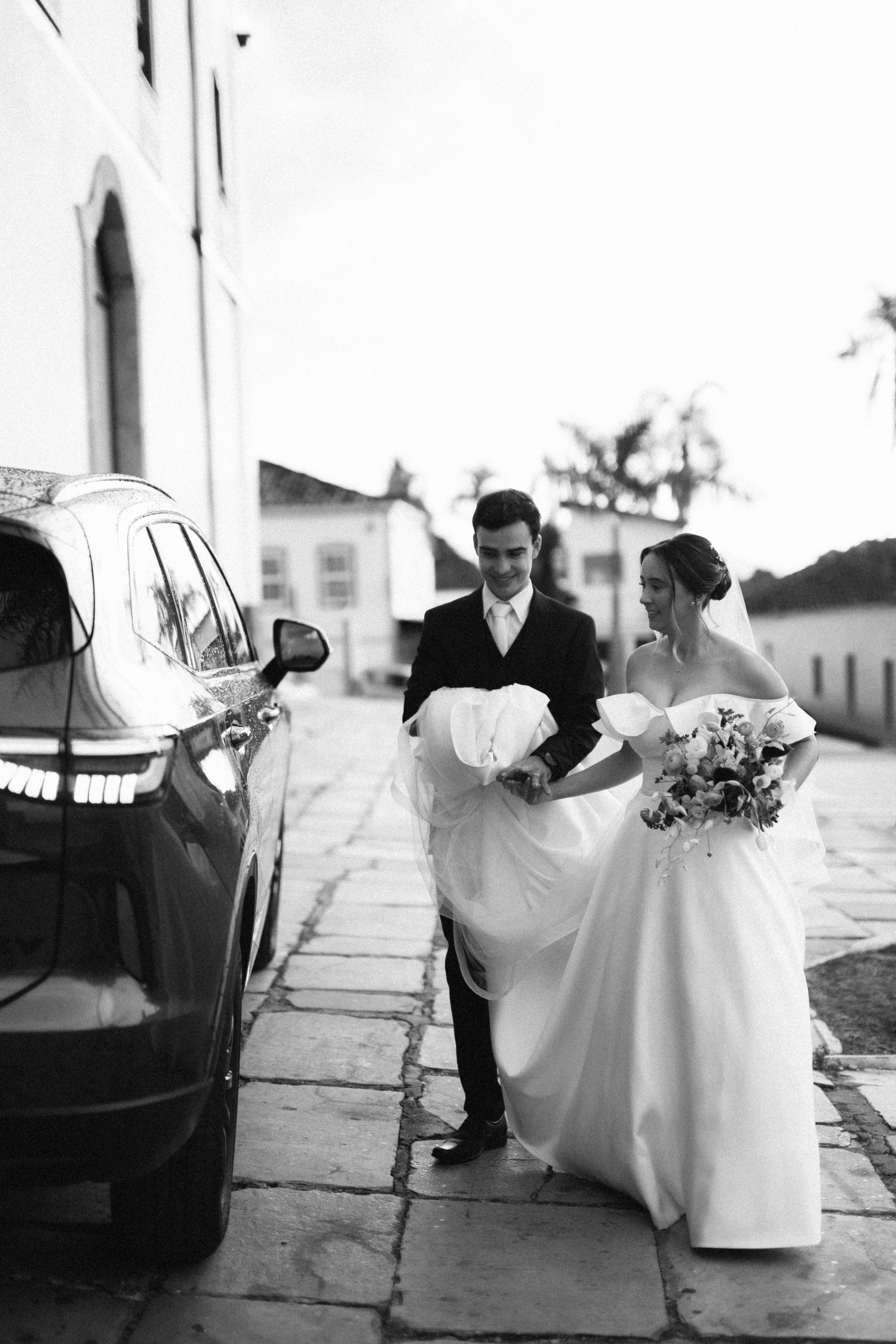Black and white photo of a bride and groom walking to their wedding car, bride holding her dress and bouquet.
