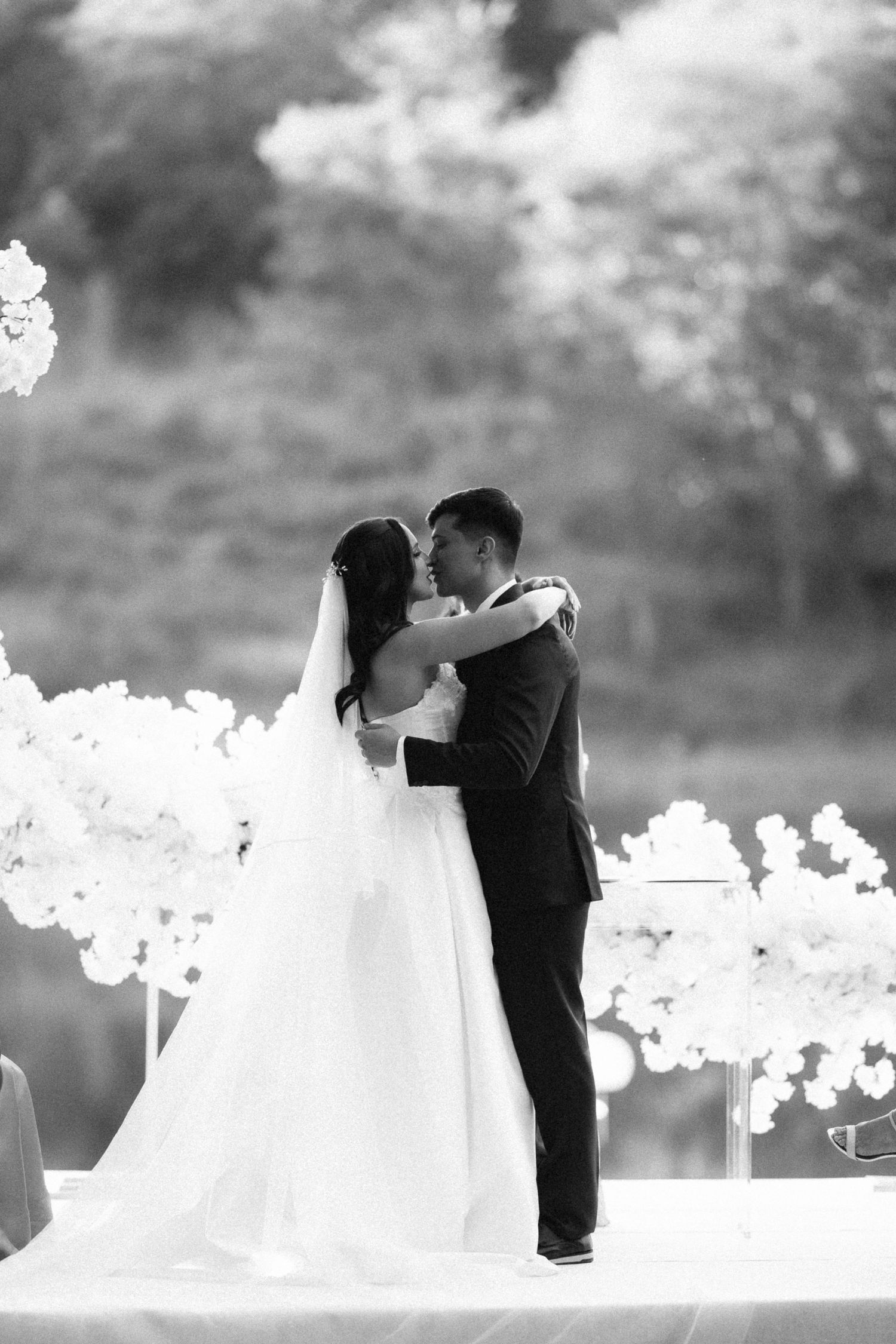 Black and white photo of a bride and groom sharing their first kiss at an outdoor wedding ceremony.