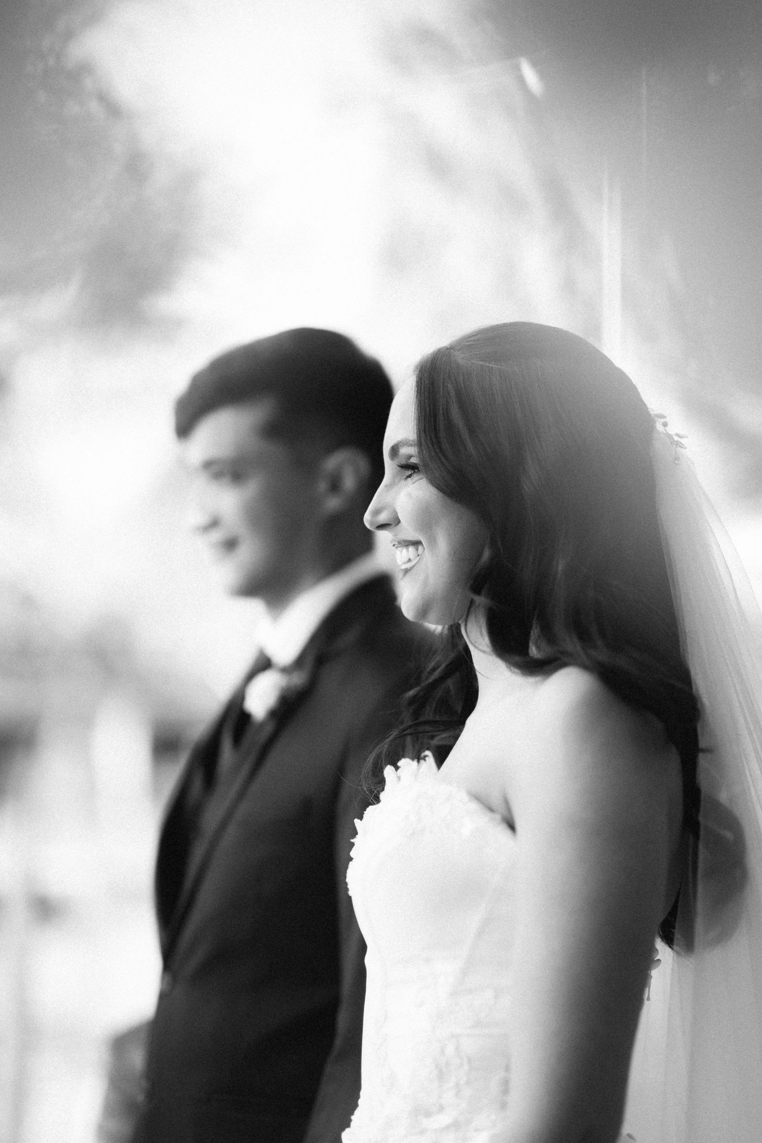 Black and white portrait of smiling bride in strapless gown with groom blurred in background.