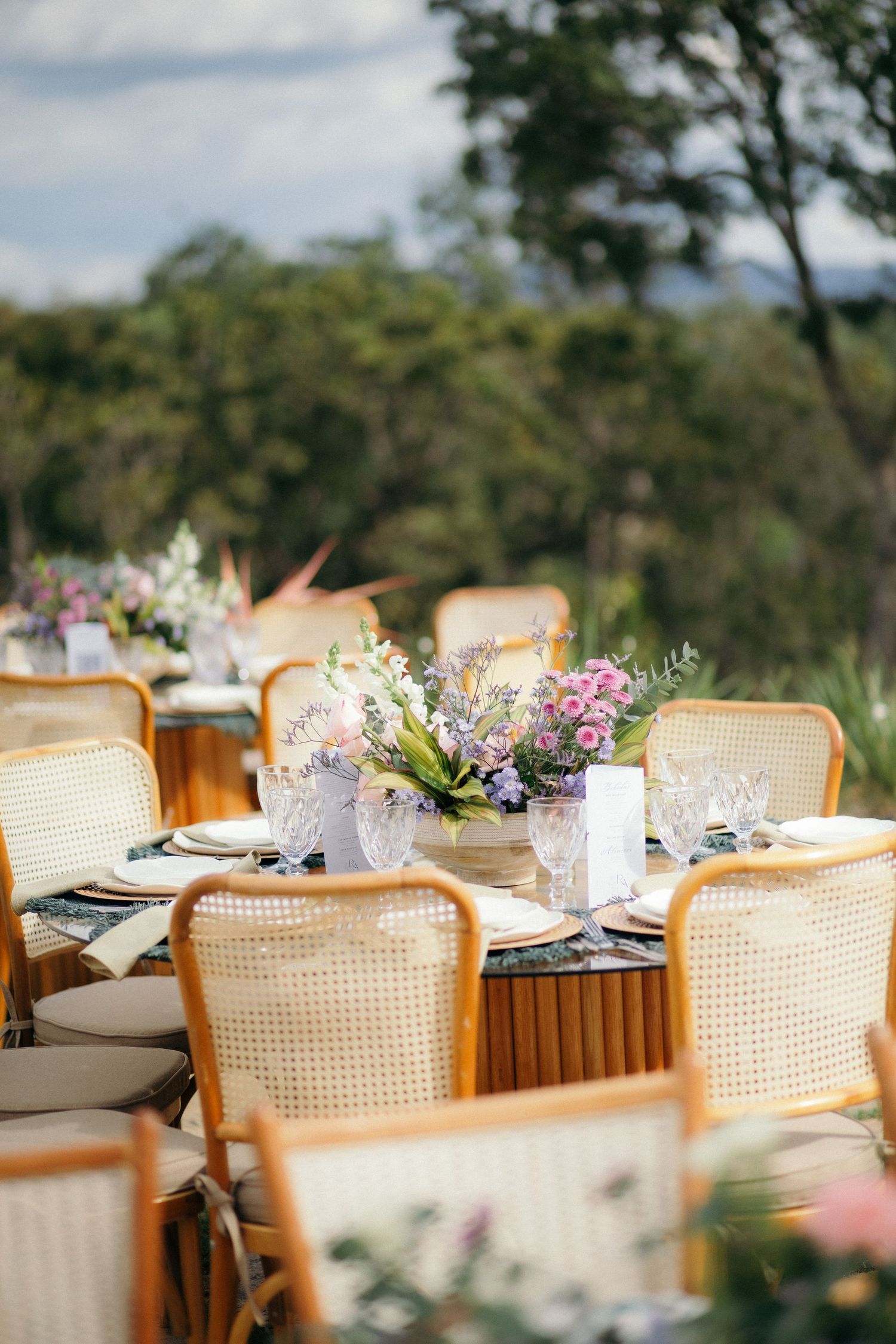 Outdoor wedding reception table with wicker chairs, floral centerpieces, and elegant place settings in a garden setting.
