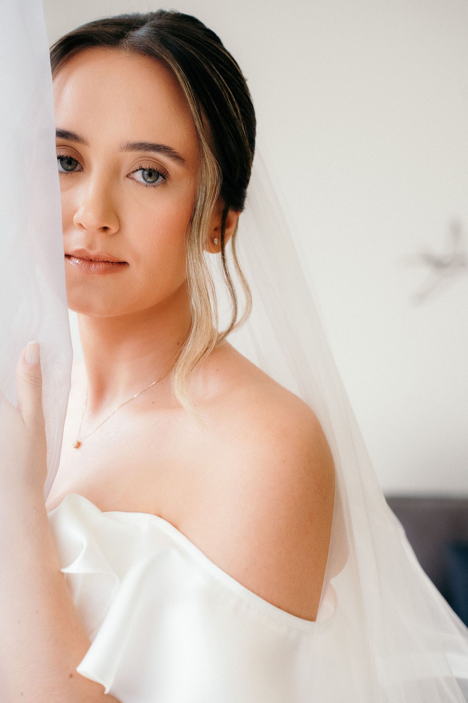 Elegant bride in off-shoulder gown with veil draped over shoulder gazing softly toward camera indoors.