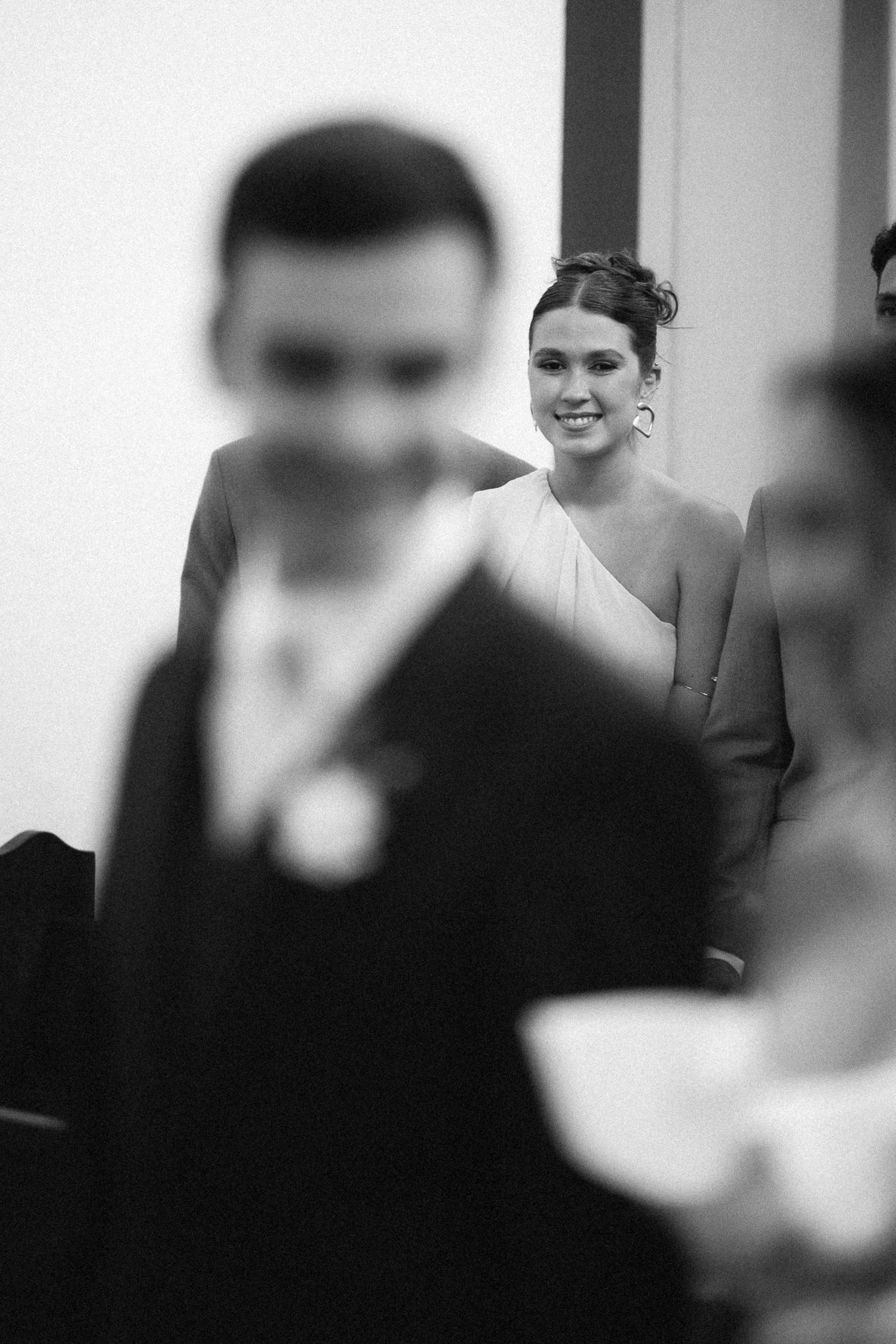 Black and white wedding photo of a smiling bridesmaid in elegant dress, with blurred guests in foreground.