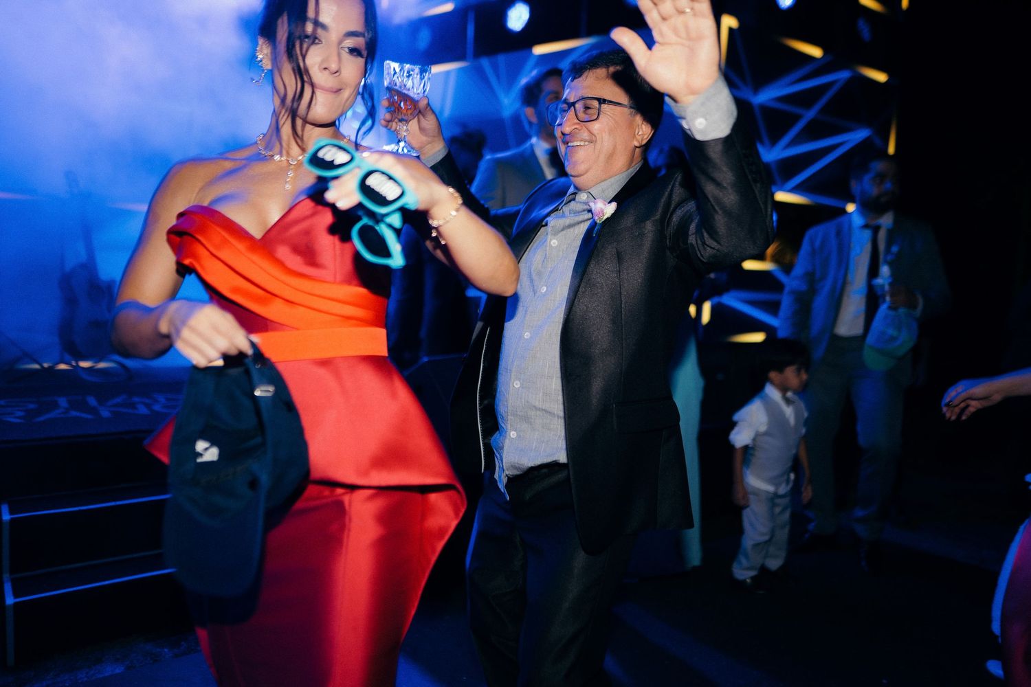 Woman in red off-shoulder gown dances energetically with older man in black jacket at vibrant nighttime wedding party.