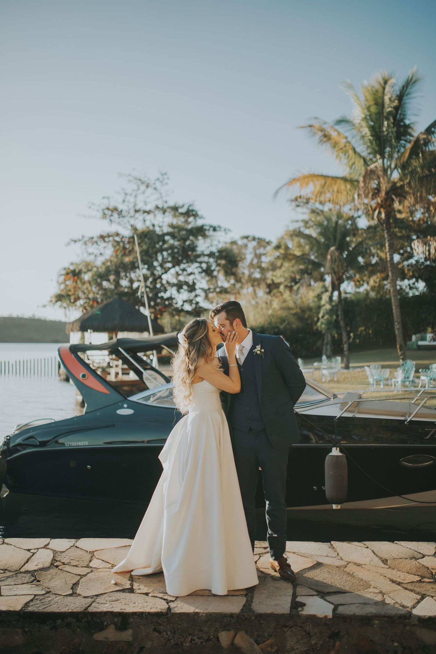 Bride and groom share a romantic kiss by a black boat near a tropical waterfront on their wedding day.
