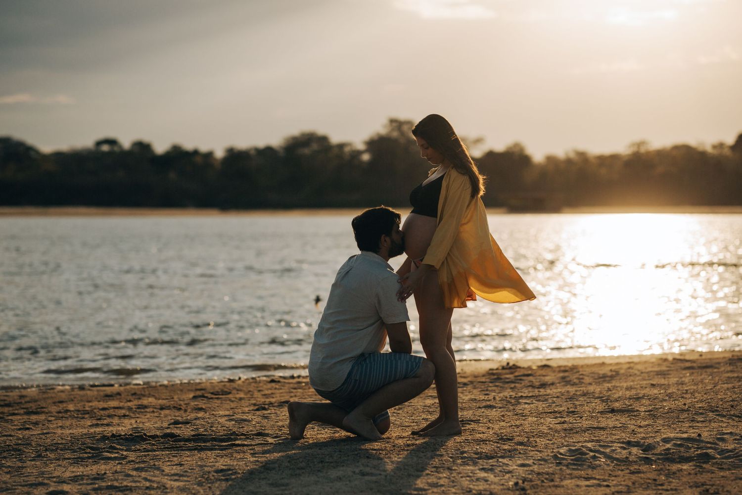 Man kneeling before pregnant woman on sandy beach at golden sunset by a lake.