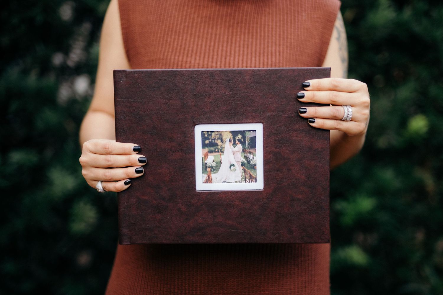 Person holding a dark brown leather wedding photo album with a polaroid picture on the cover, outdoors.