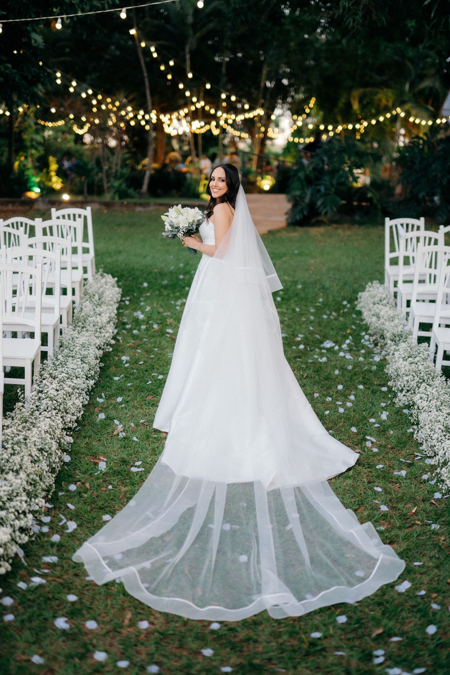 Bride in elegant white ball gown with long veil walks down outdoor aisle lined with white chairs and floral arrangements.