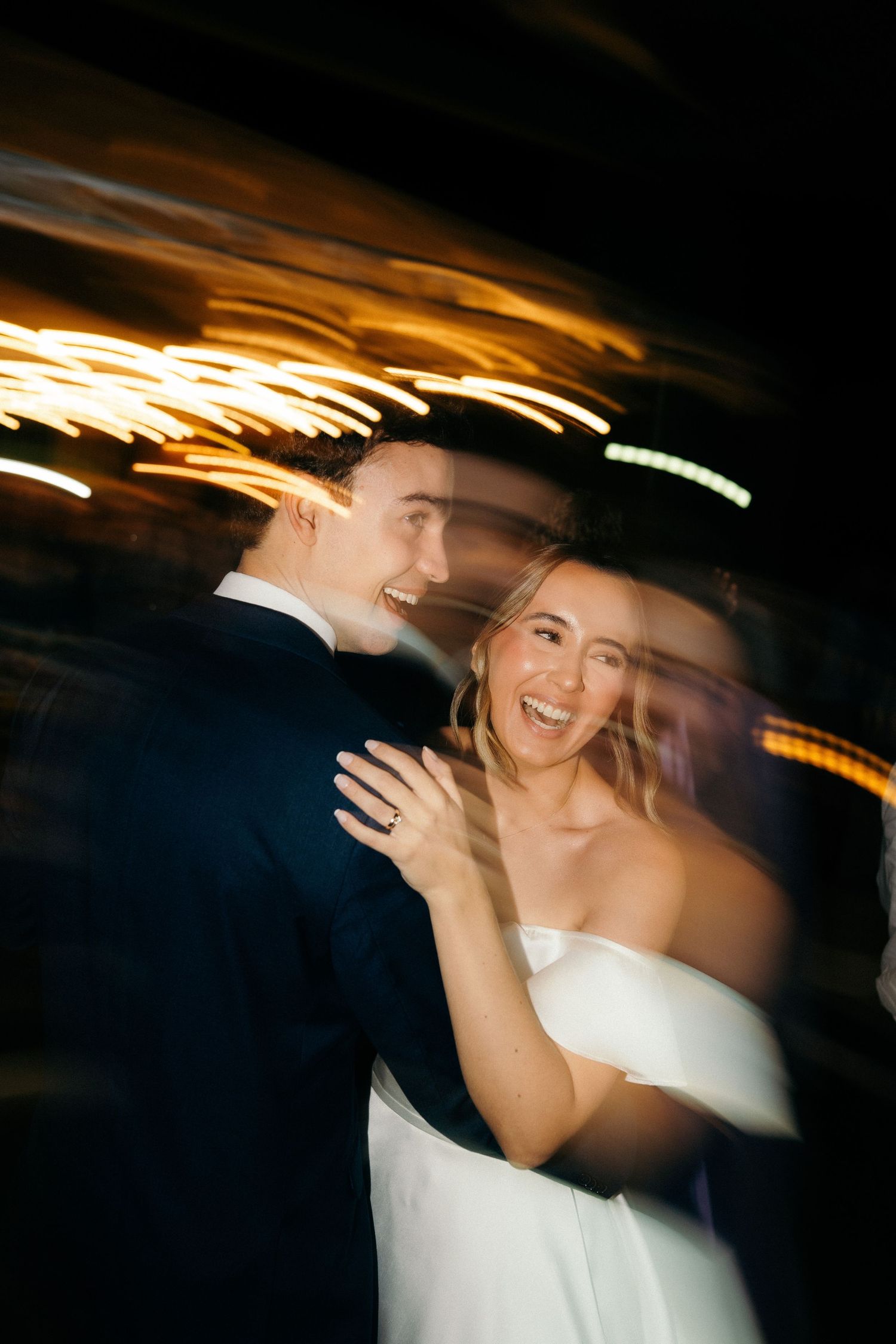 Motion blur photo of a joyful bride and groom dancing together under golden lights at their wedding reception.