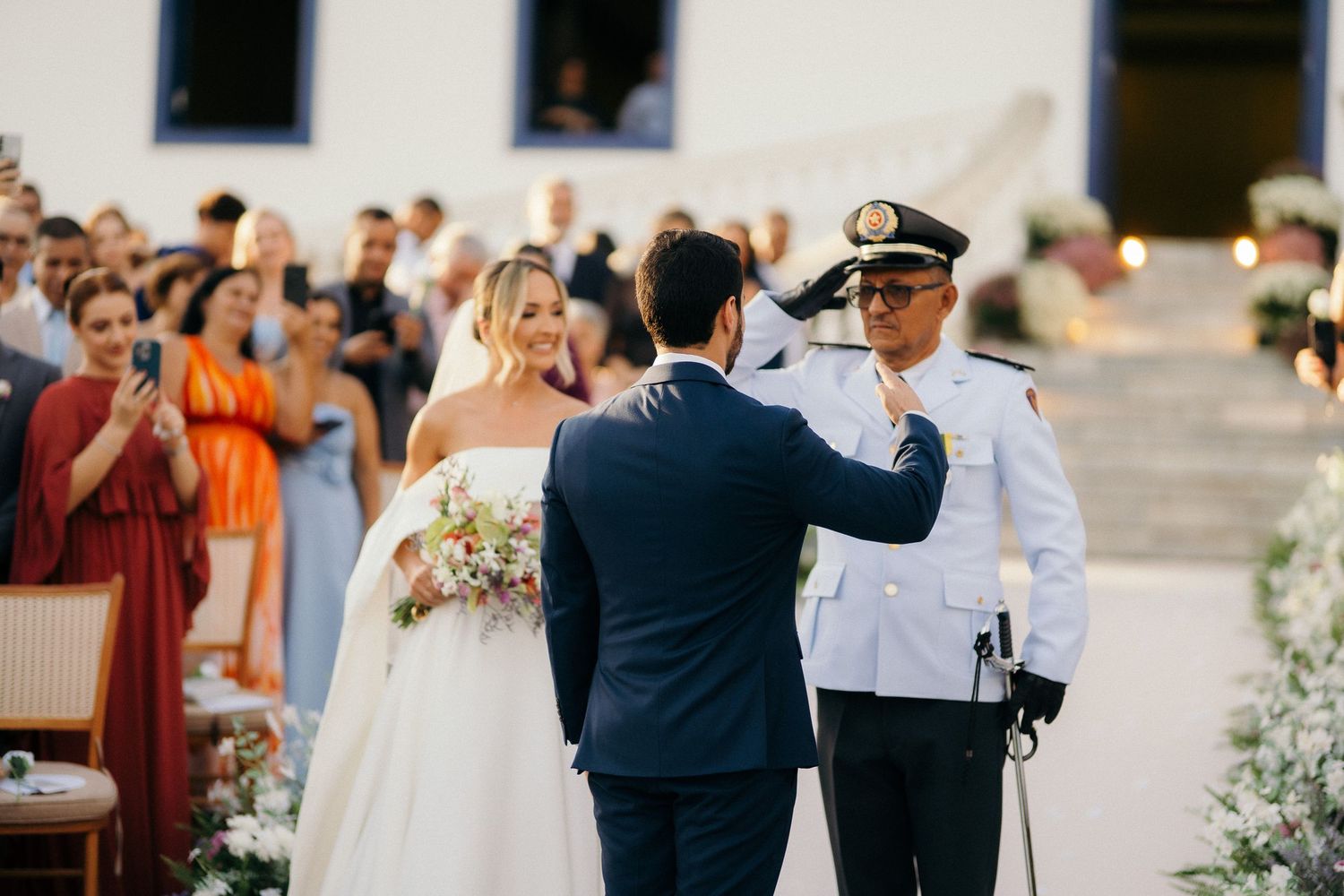 Military officer salutes bride and groom during outdoor wedding ceremony with guests watching.