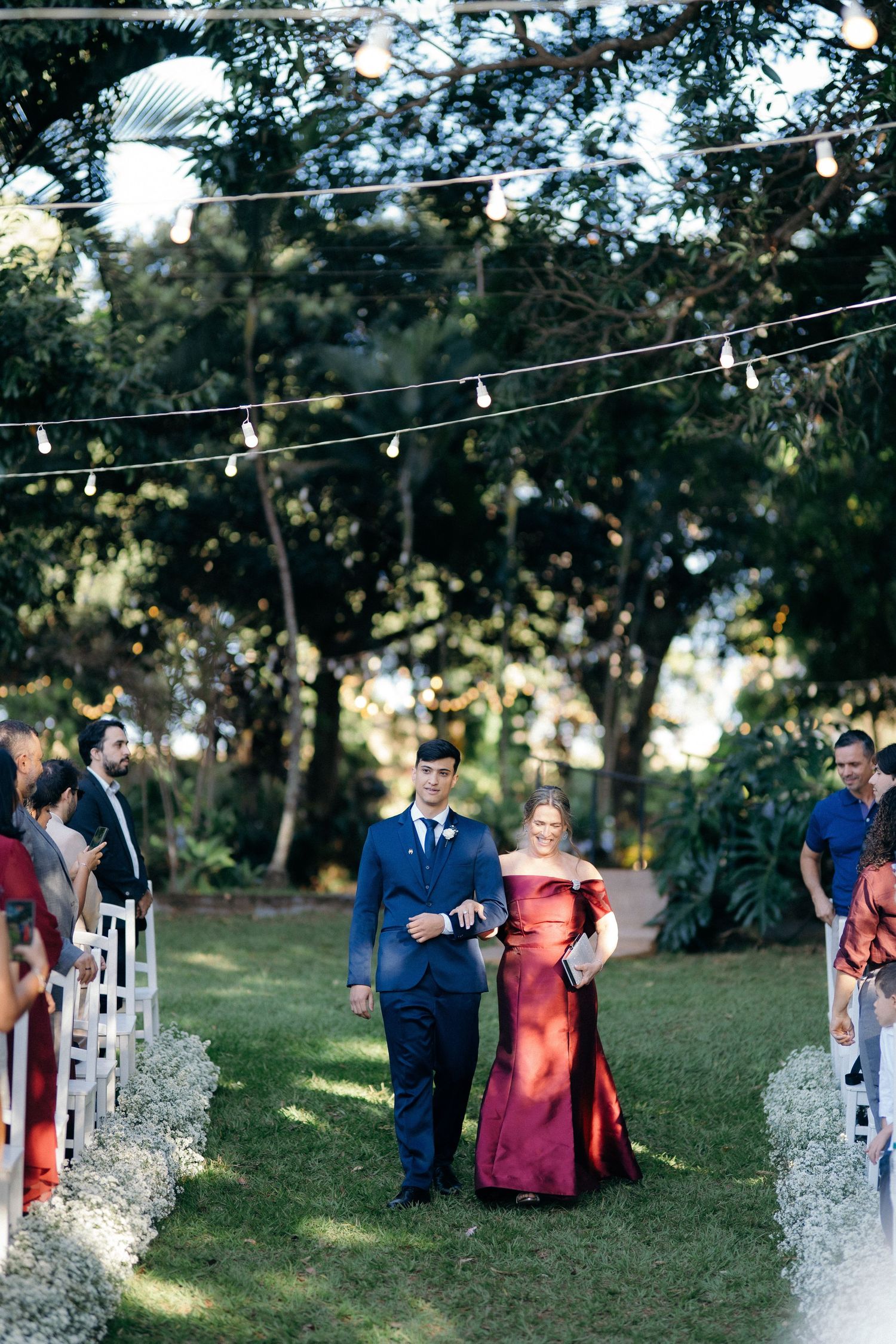 Groom in blue suit walks down outdoor wedding aisle with woman in red gown under string lights.
