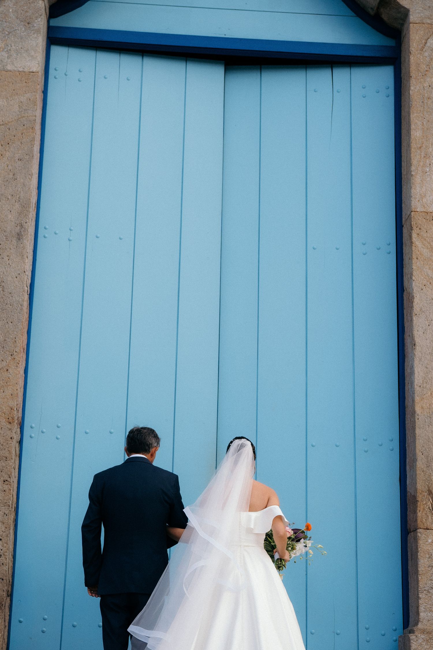 Bride and groom standing before a large blue wooden door, bride holding a bouquet in a white gown with veil.