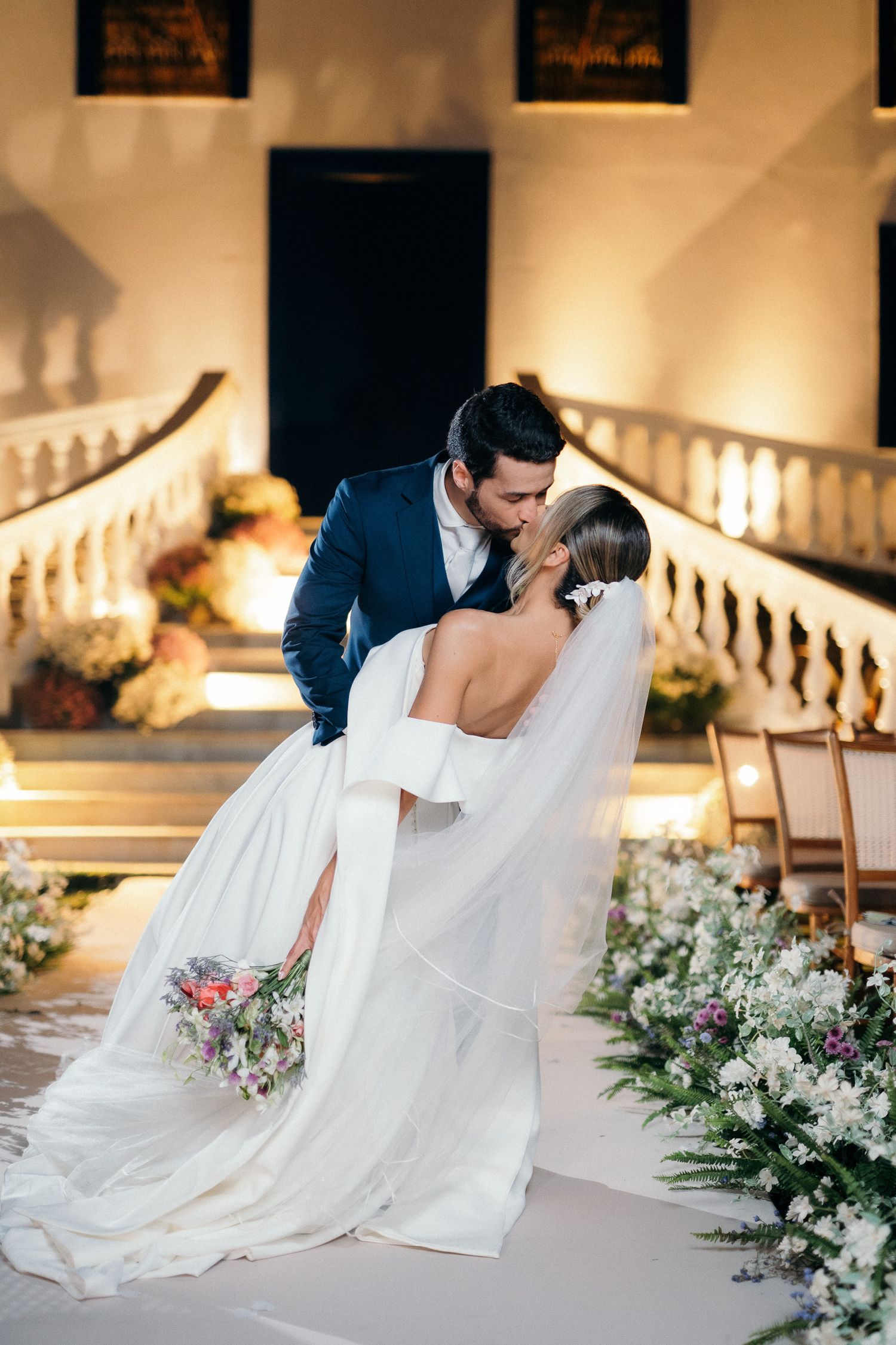 Groom dips bride for a kiss at elegant evening wedding ceremony with grand staircase backdrop.