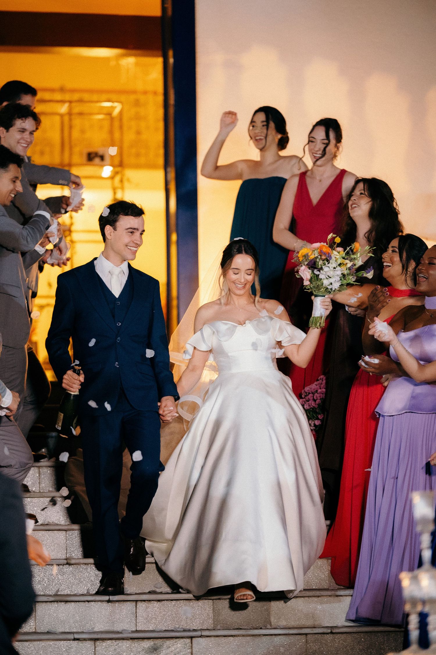 Smiling newlyweds descend steps surrounded by cheering bridesmaids in colorful gowns after wedding ceremony.
