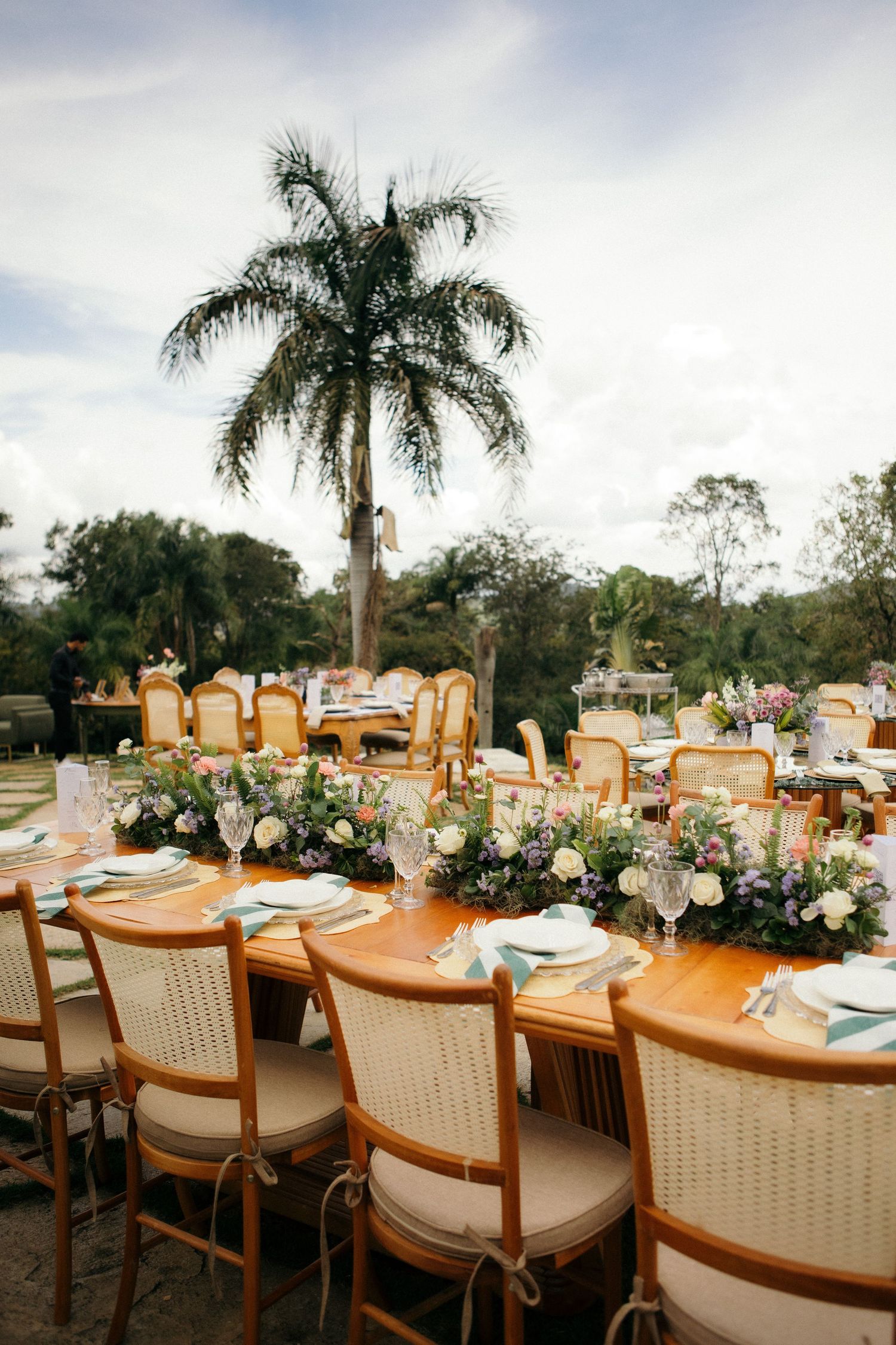 Elegant outdoor wedding reception with rattan chairs, floral centerpieces, and a tropical palm tree backdrop.