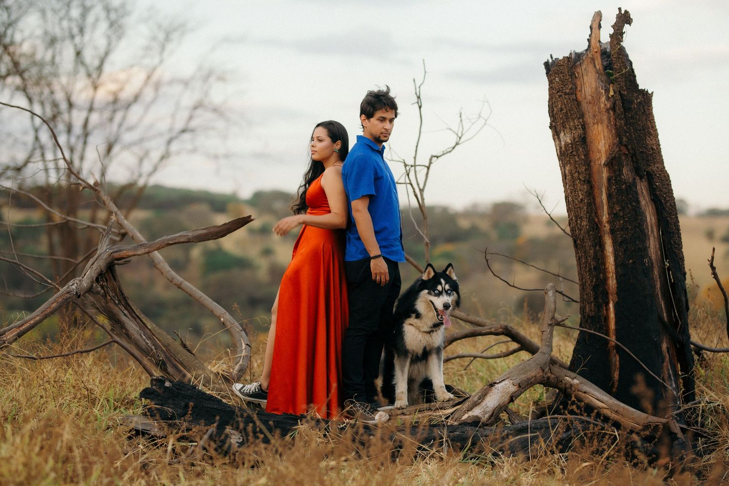 Couple standing back to back with a husky dog in a dry outdoor field.