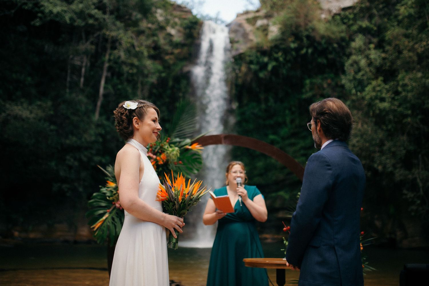 Bride laughs joyfully holding tropical bouquet during outdoor waterfall wedding ceremony with officiant.