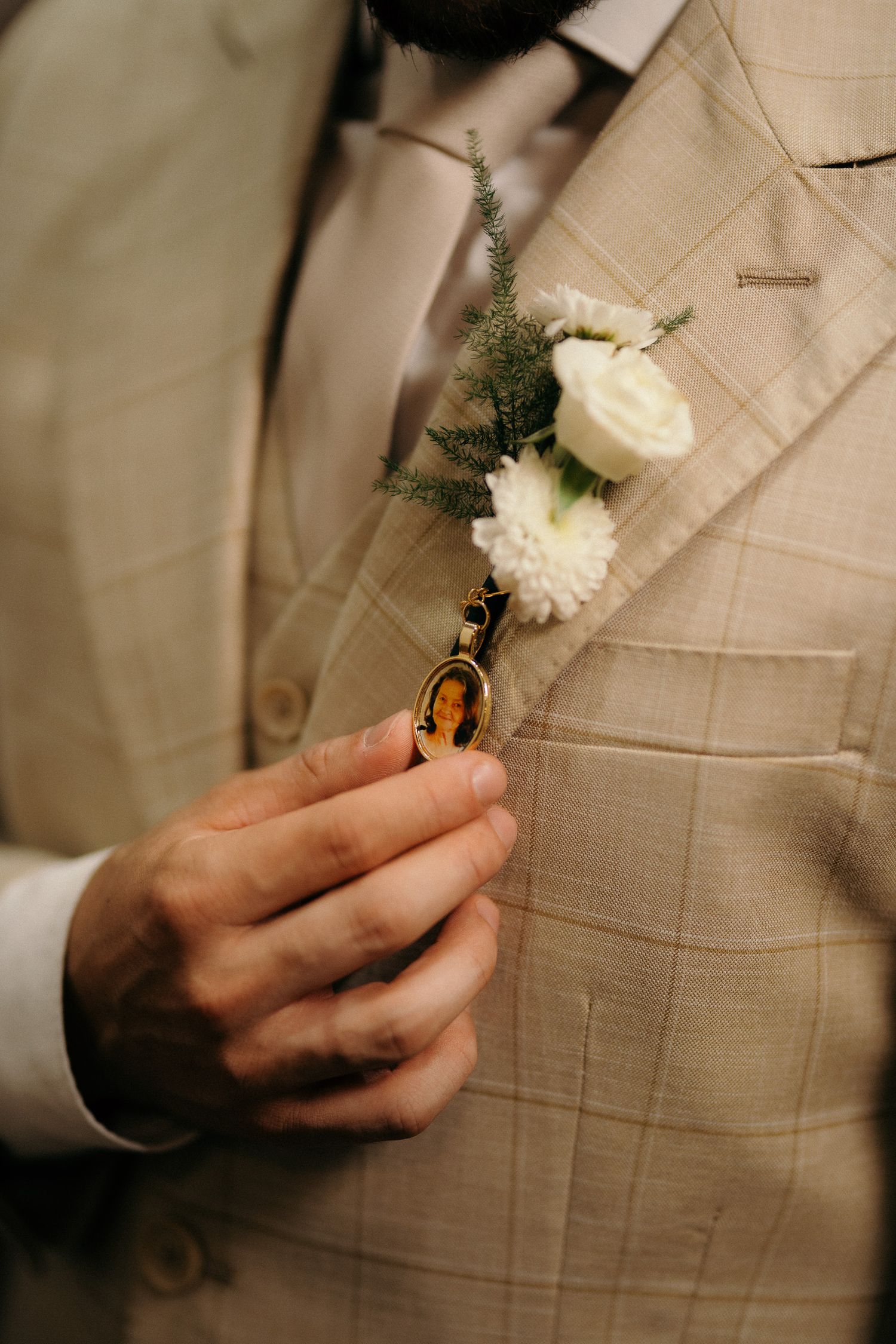 Close-up of groom holding gold locket beside white rose boutonniere on beige checkered suit lapel.
