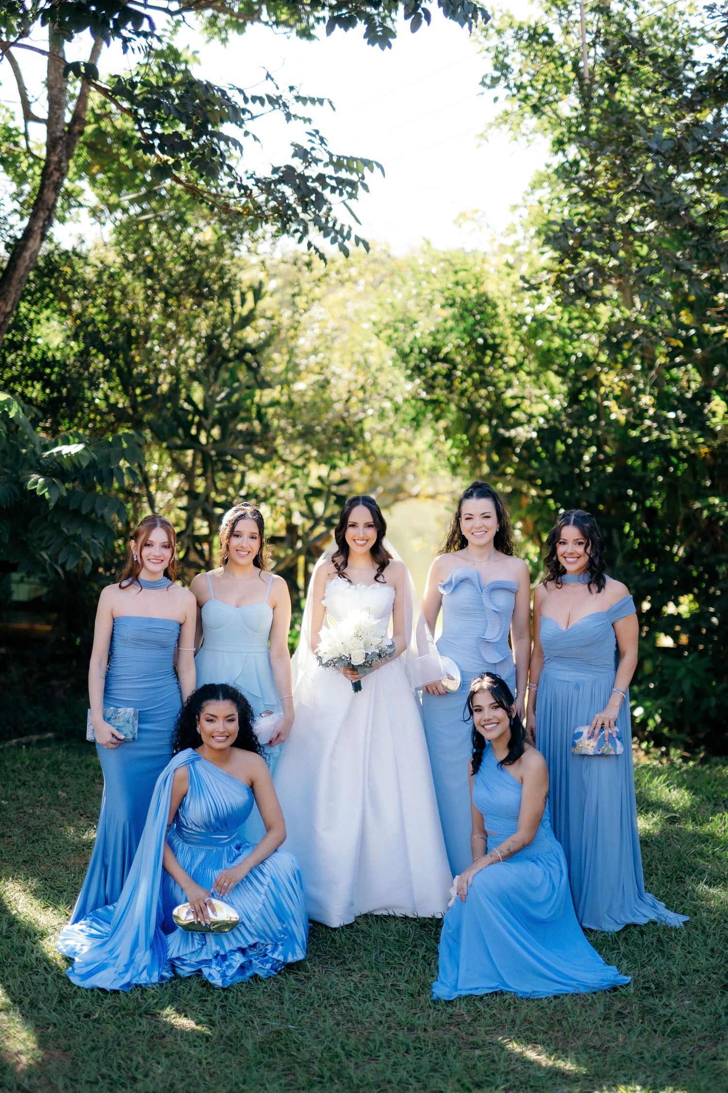 Bride in white gown with six bridesmaids in blue dresses posing outdoors in a garden.