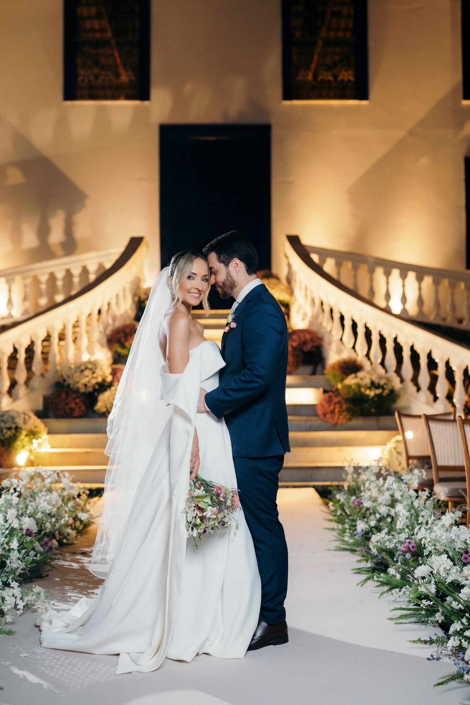 Bride and groom share romantic moment on illuminated outdoor aisle flanked by white florals.