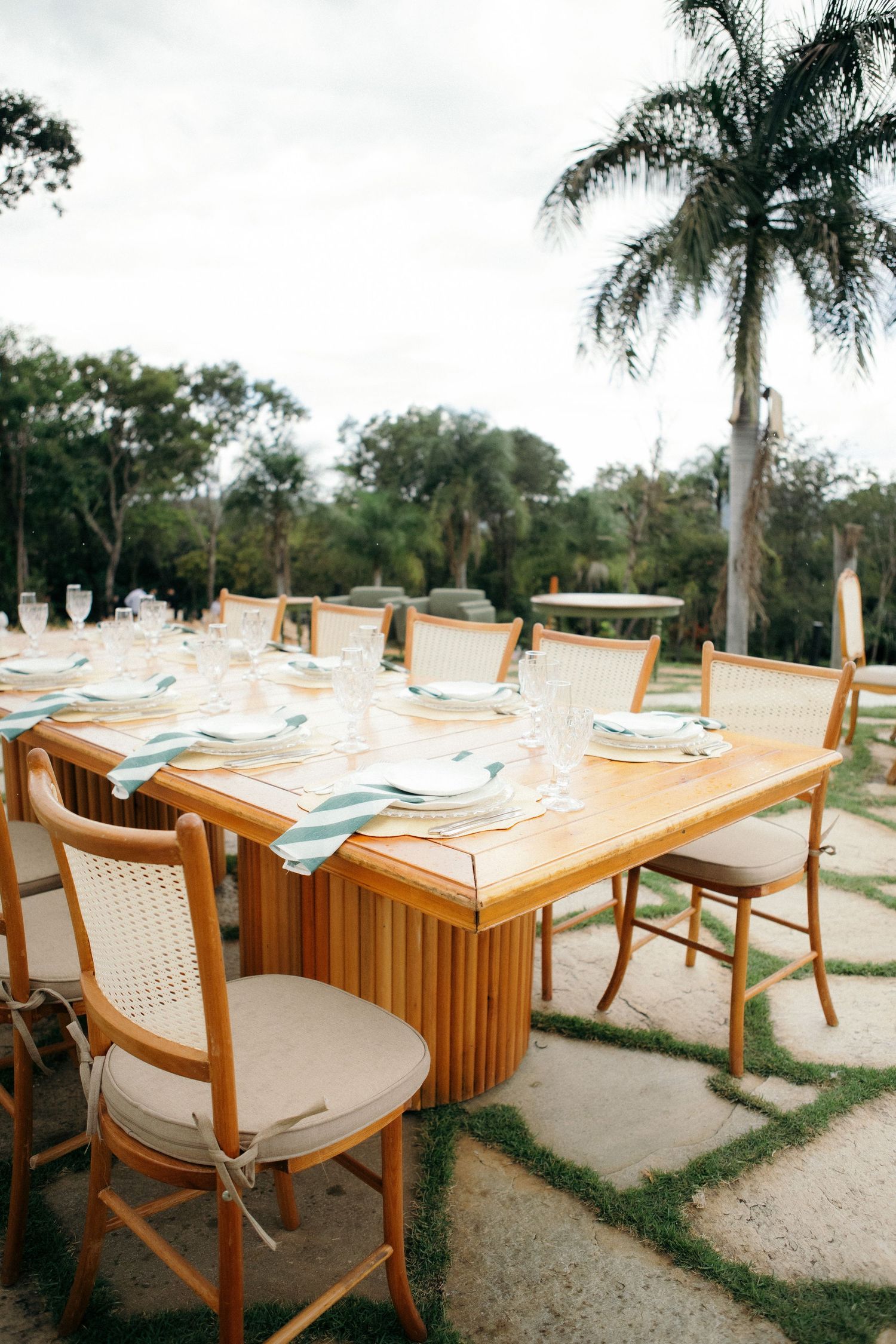 Elegant outdoor dining table with wooden chairs, light blue napkins, and glassware set against lush palm trees.