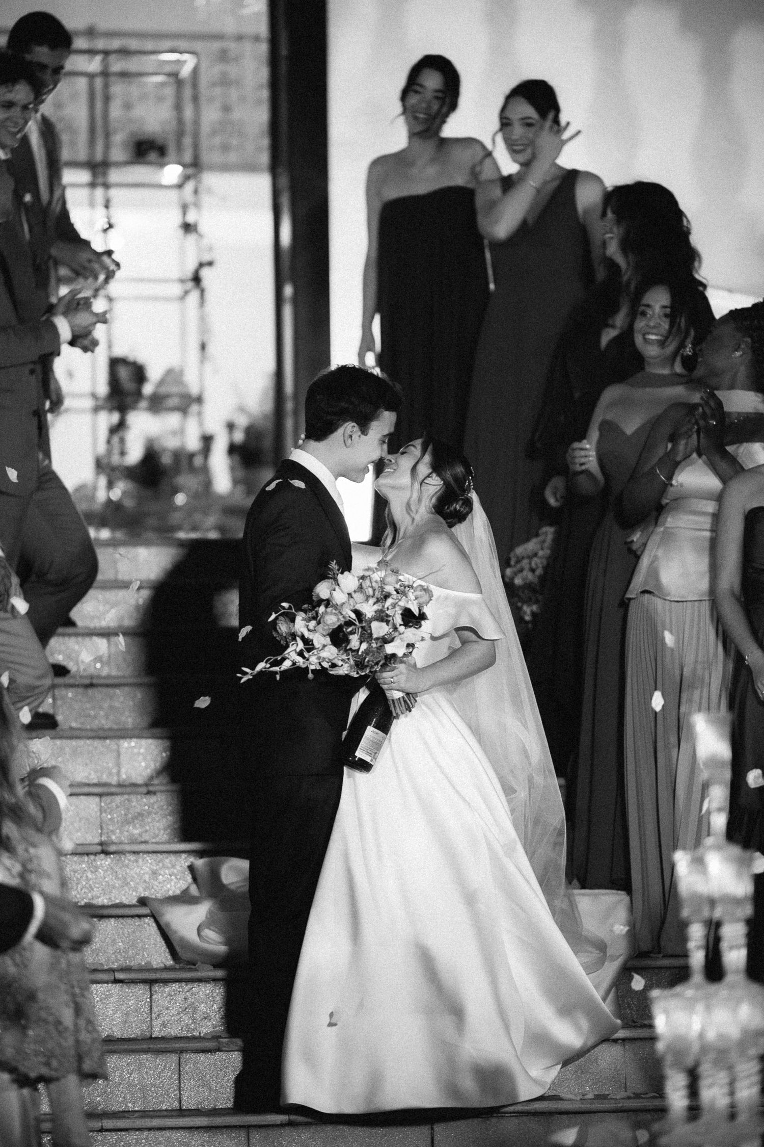 Black and white photo of bride and groom kissing on stairs surrounded by bridesmaids at elegant wedding ceremony.