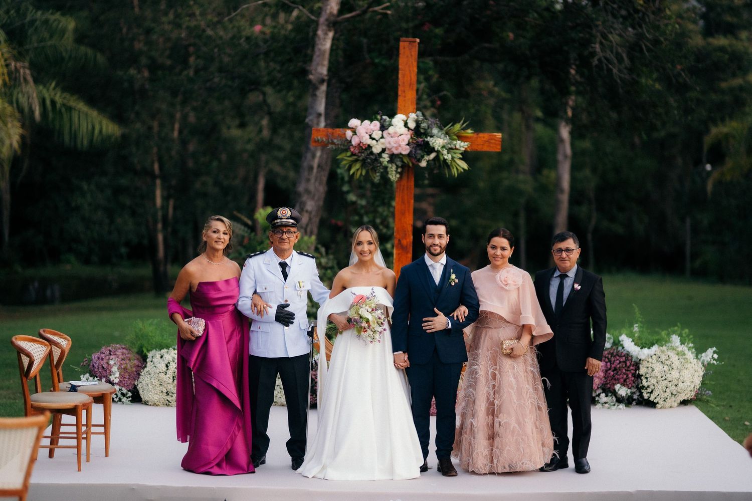 Wedding party poses before wooden cross altar with floral arrangements at outdoor garden ceremony.