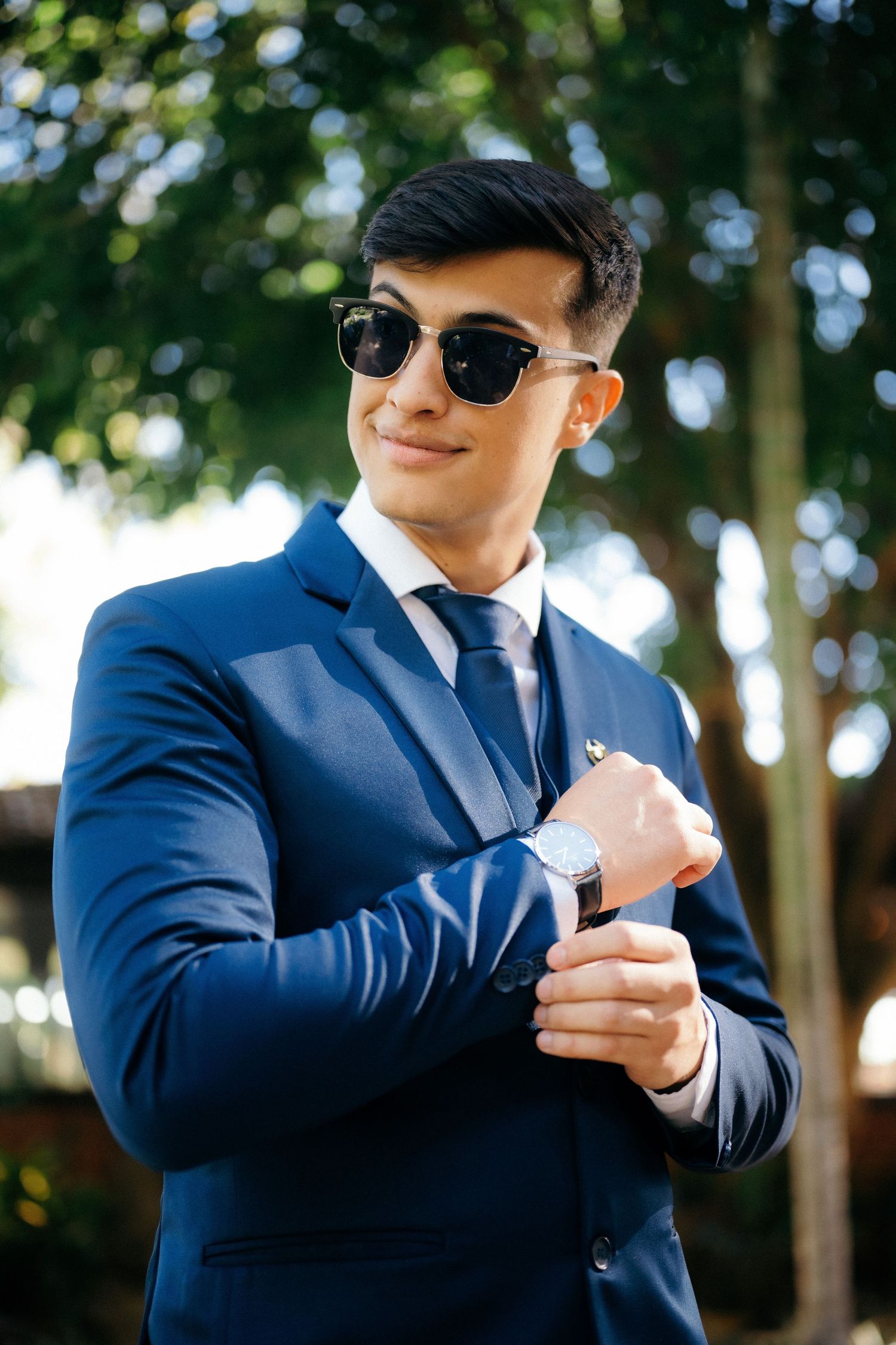 Stylish young man in navy blue suit and sunglasses checking his watch outdoors with trees in background.