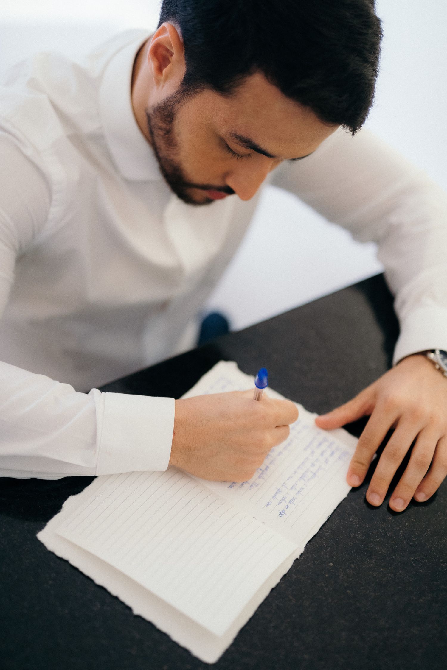 A bearded businessman in a white shirt writing notes in a lined notebook with a blue pen at a dark desk.