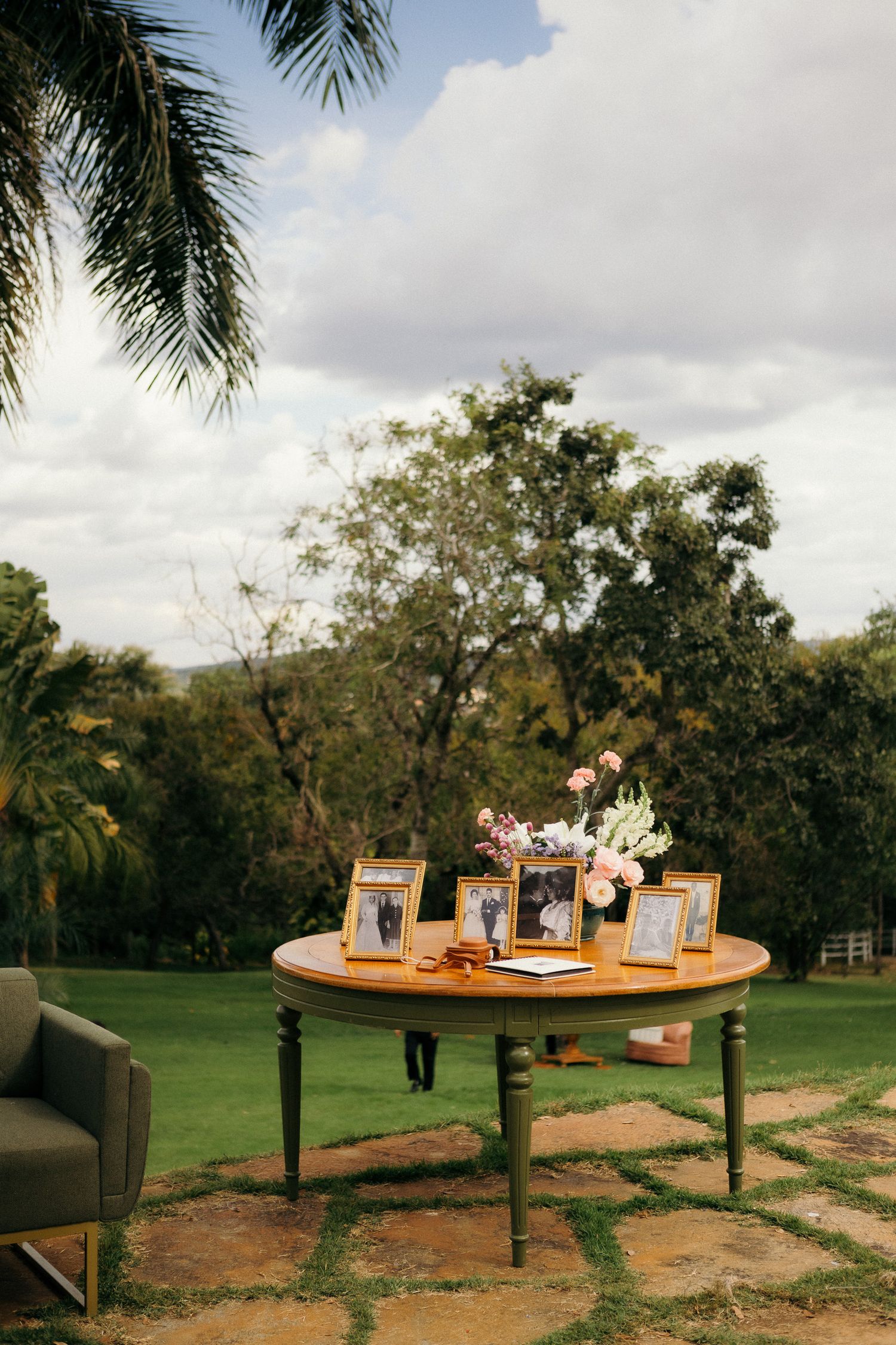 Elegant outdoor wedding table with gold frames, floral arrangement, and guest book on lush green lawn.