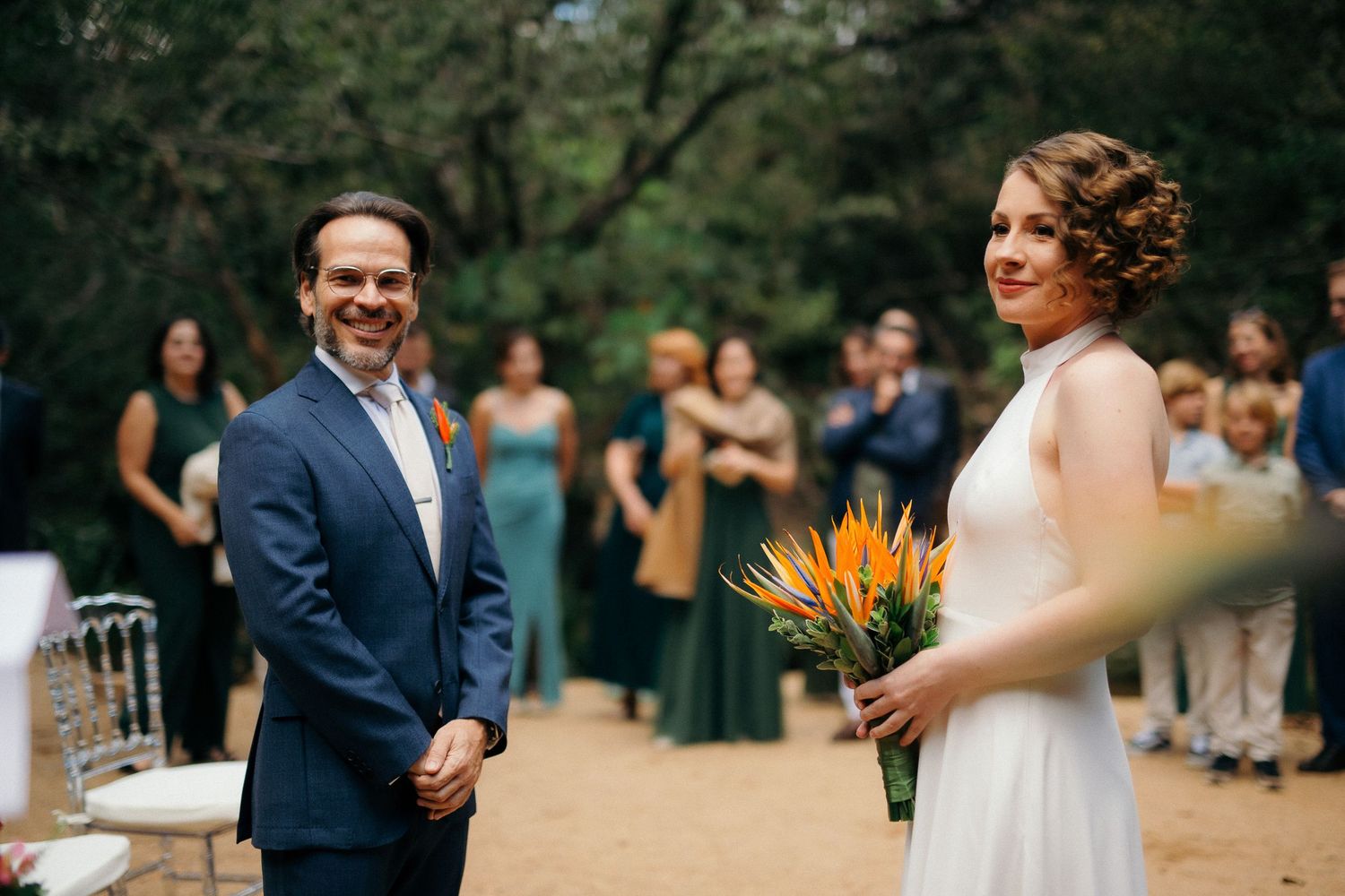 Bride holding vibrant bird of paradise bouquet smiles at groom during an outdoor woodland wedding ceremony.