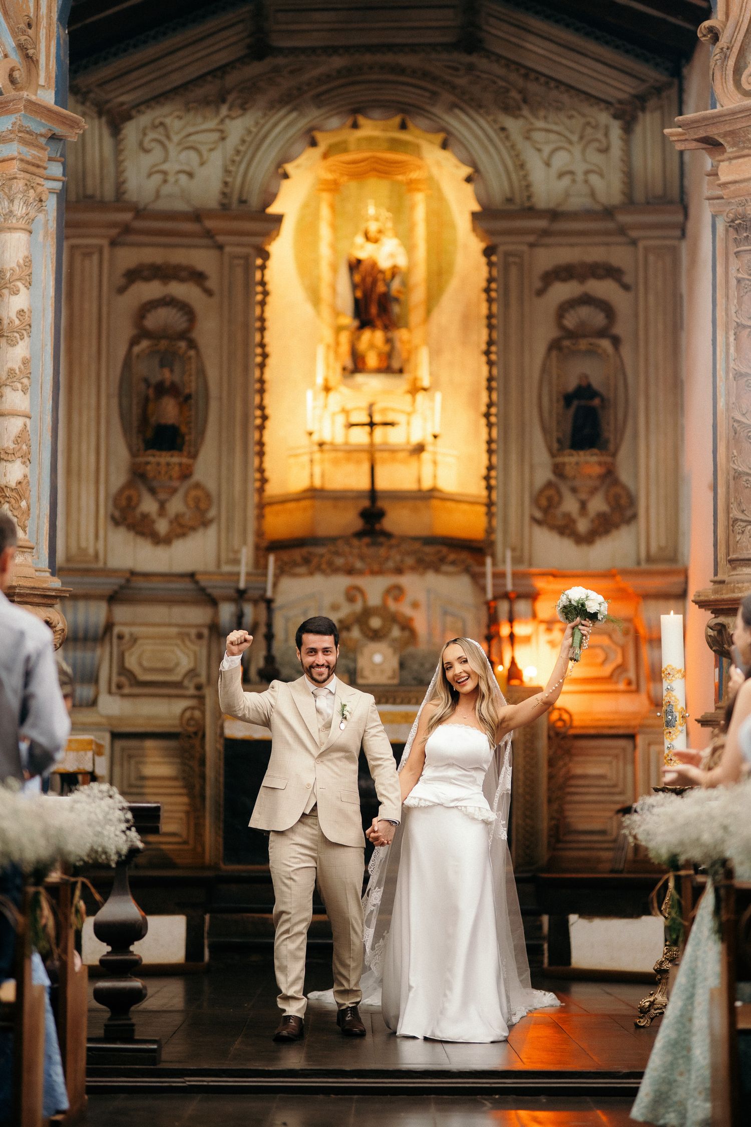 Joyful newlyweds celebrate at church altar, bride holding bouquet aloft in elegant white gown, groom in beige suit.