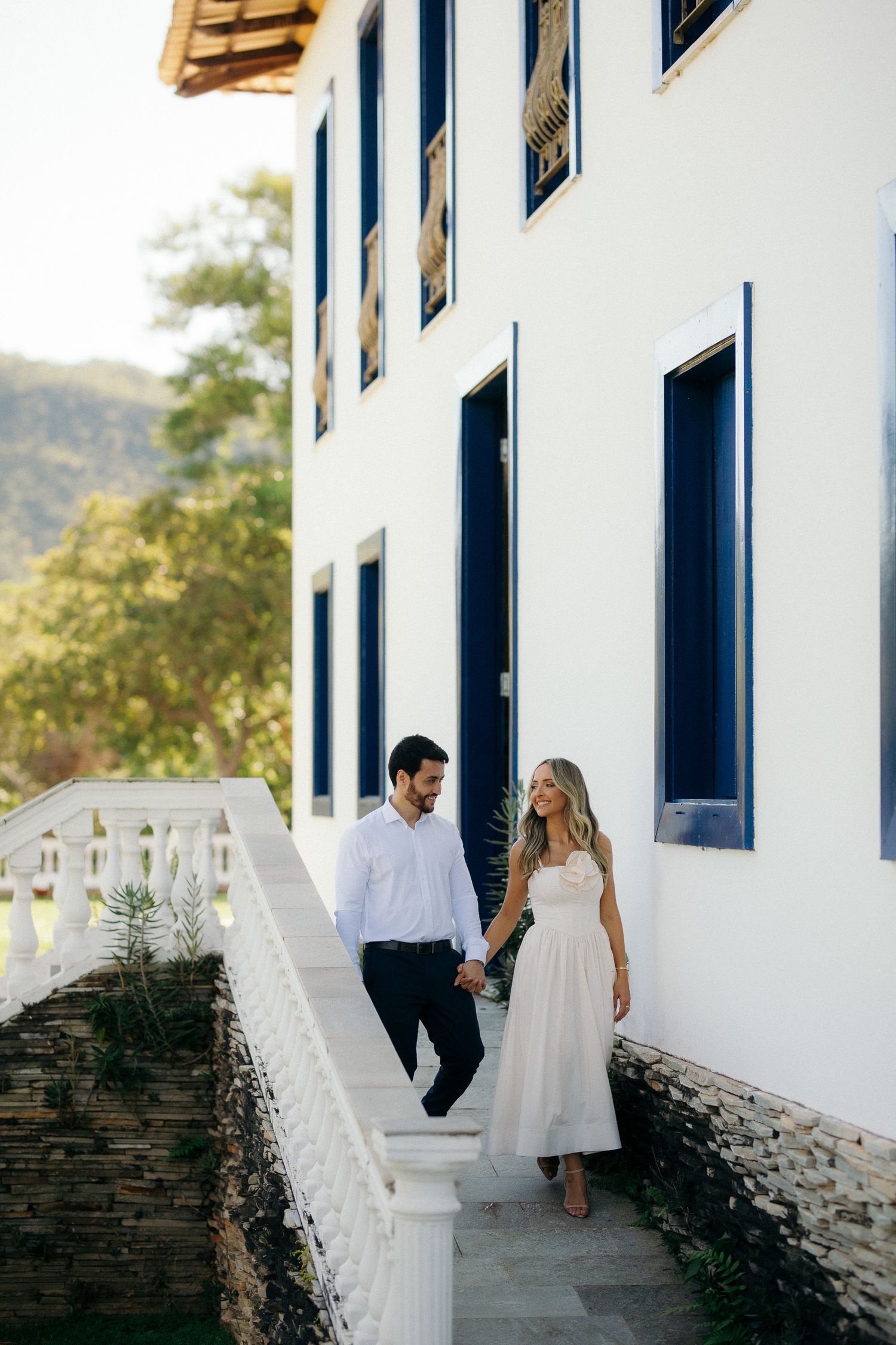 Couple holding hands walking down steps beside white colonial building with blue shutters and mountains.