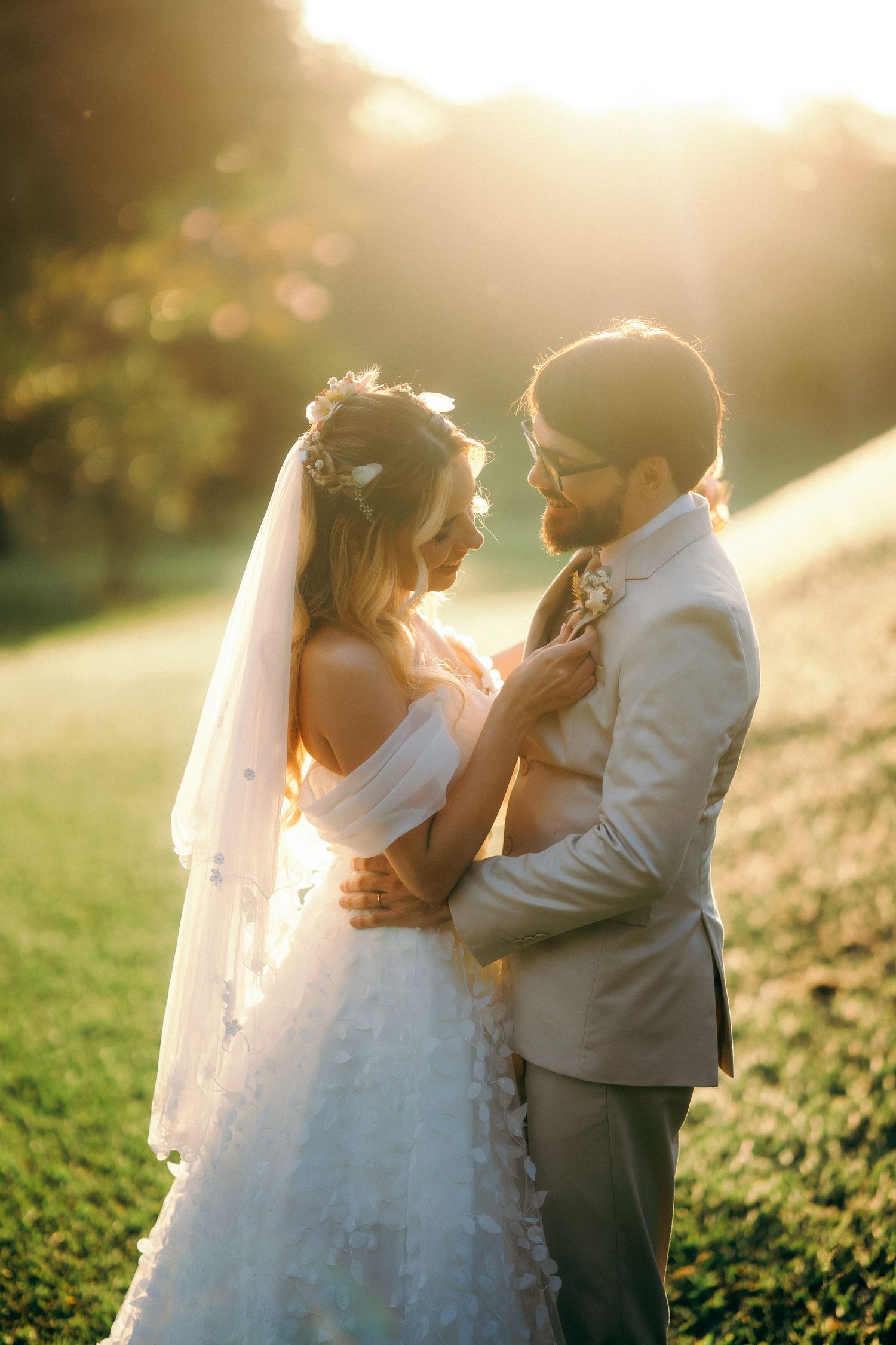 Bride and groom embrace in golden sunset light on green lawn, she wears white gown and veil, he wears gray suit.