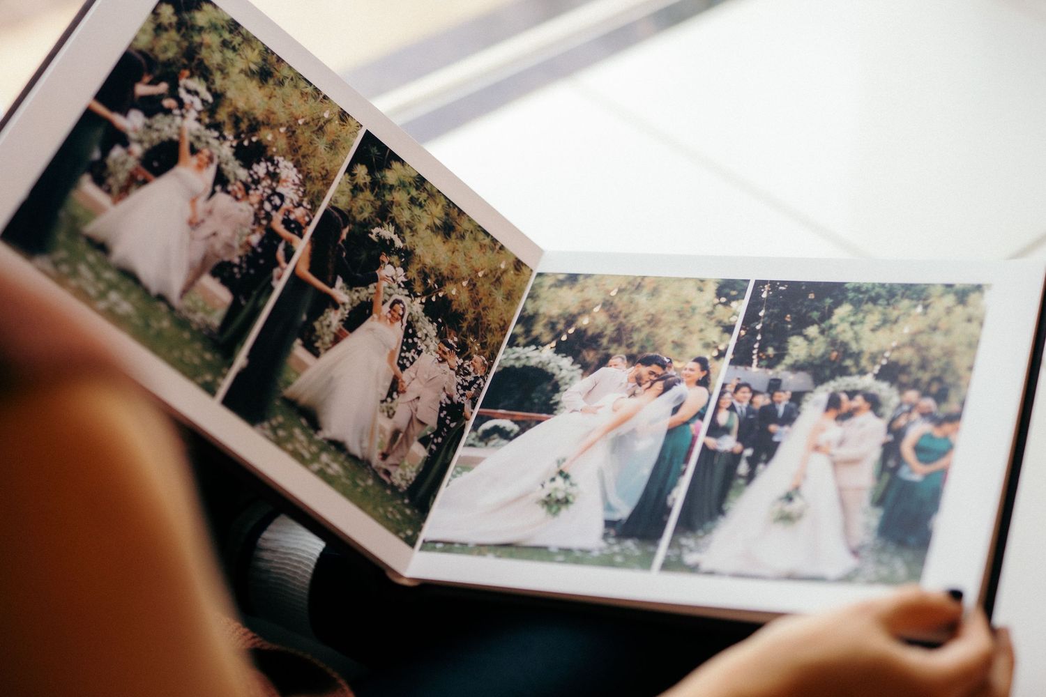 Person holding open a wedding photo album showing bride and groom celebration photos in an outdoor ceremony.