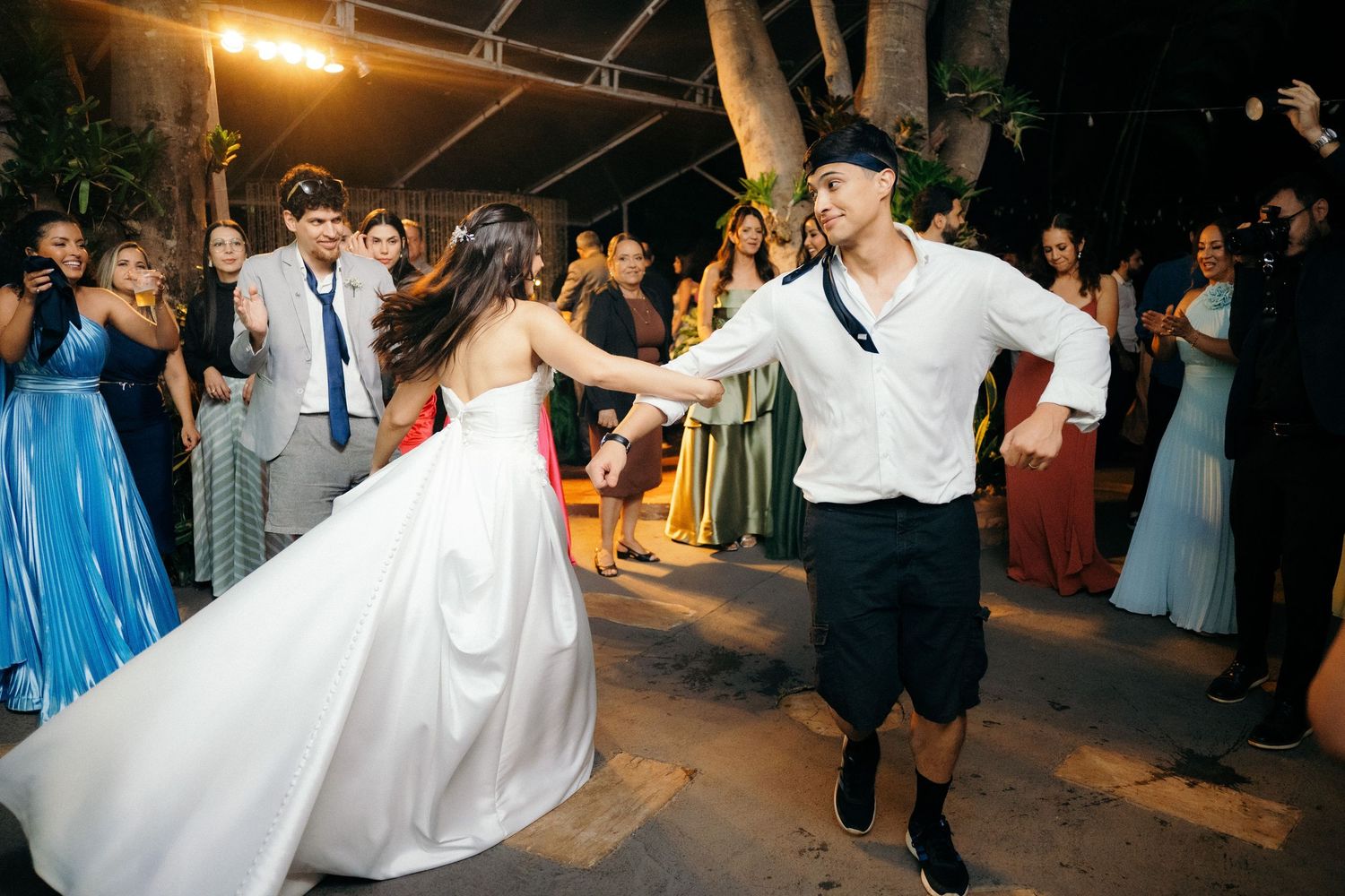 Bride in white gown dancing with casually dressed guest at lively outdoor wedding reception.